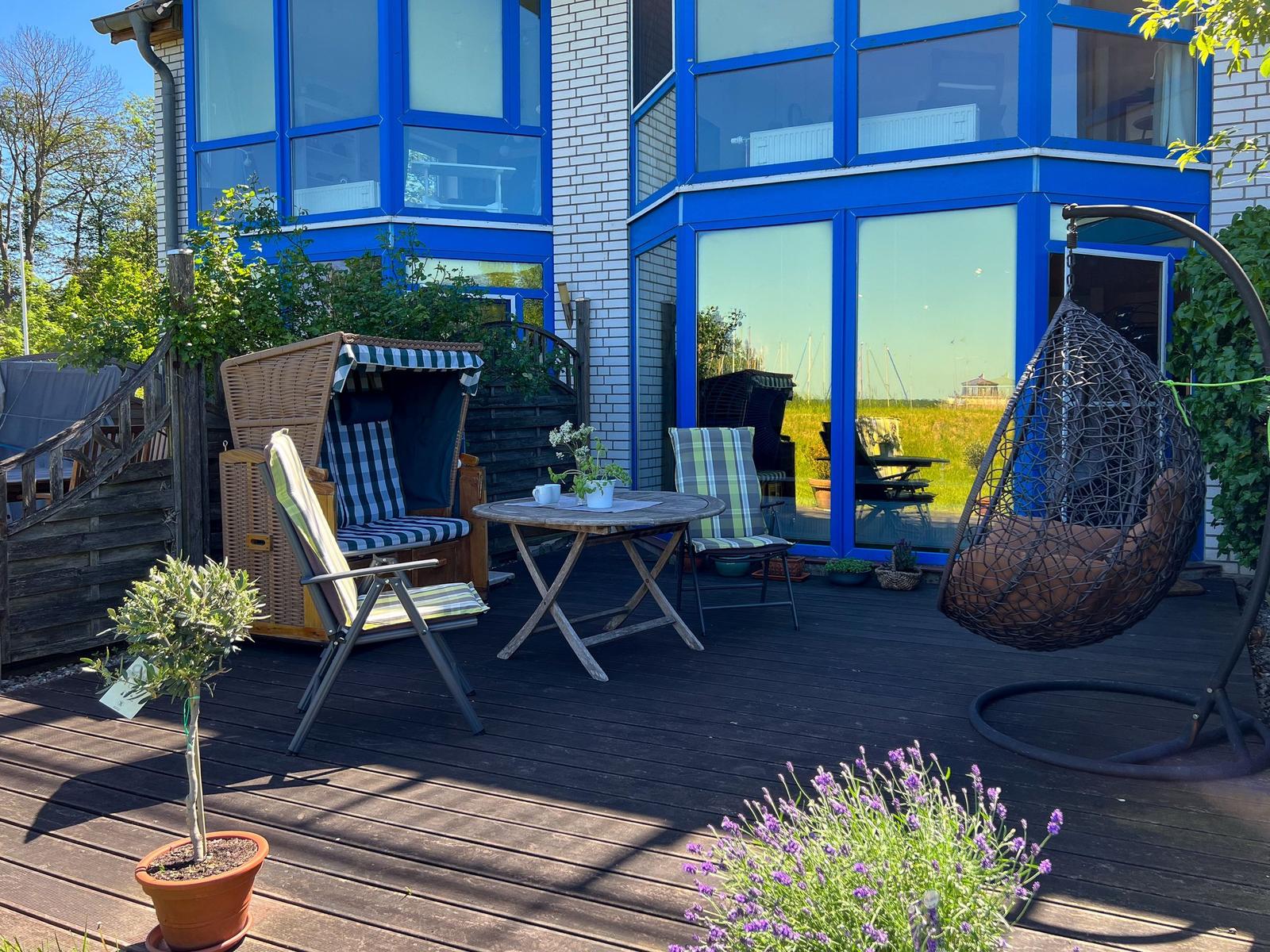 Terrasse mit Sitzgelegenheiten und Pflanzen vor einem Haus mit blauen Fensterrahmen.