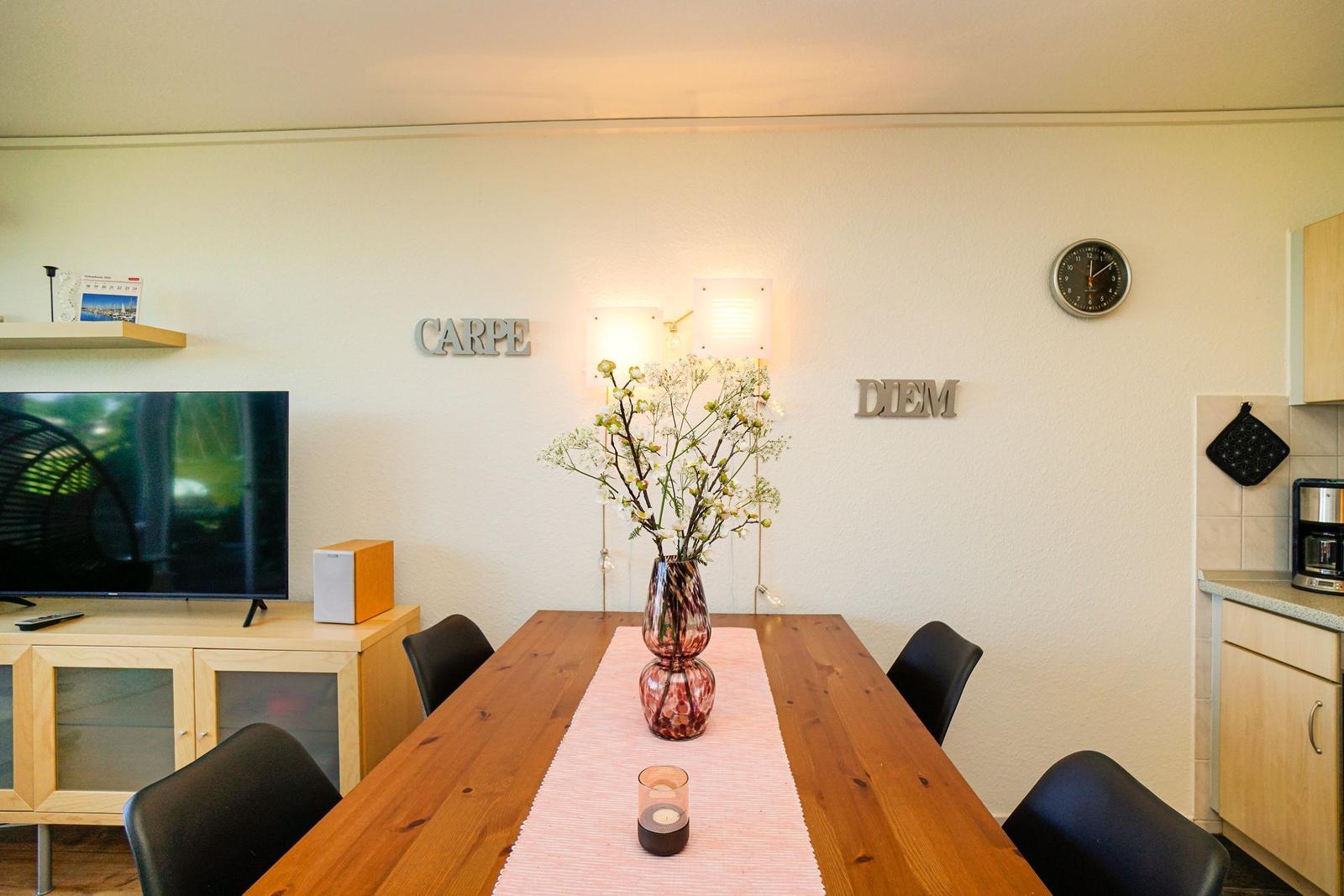 Esszimmer mit Holztisch, Stühlen und Blumenvase. Wanddekoration und Uhr sichtbar.