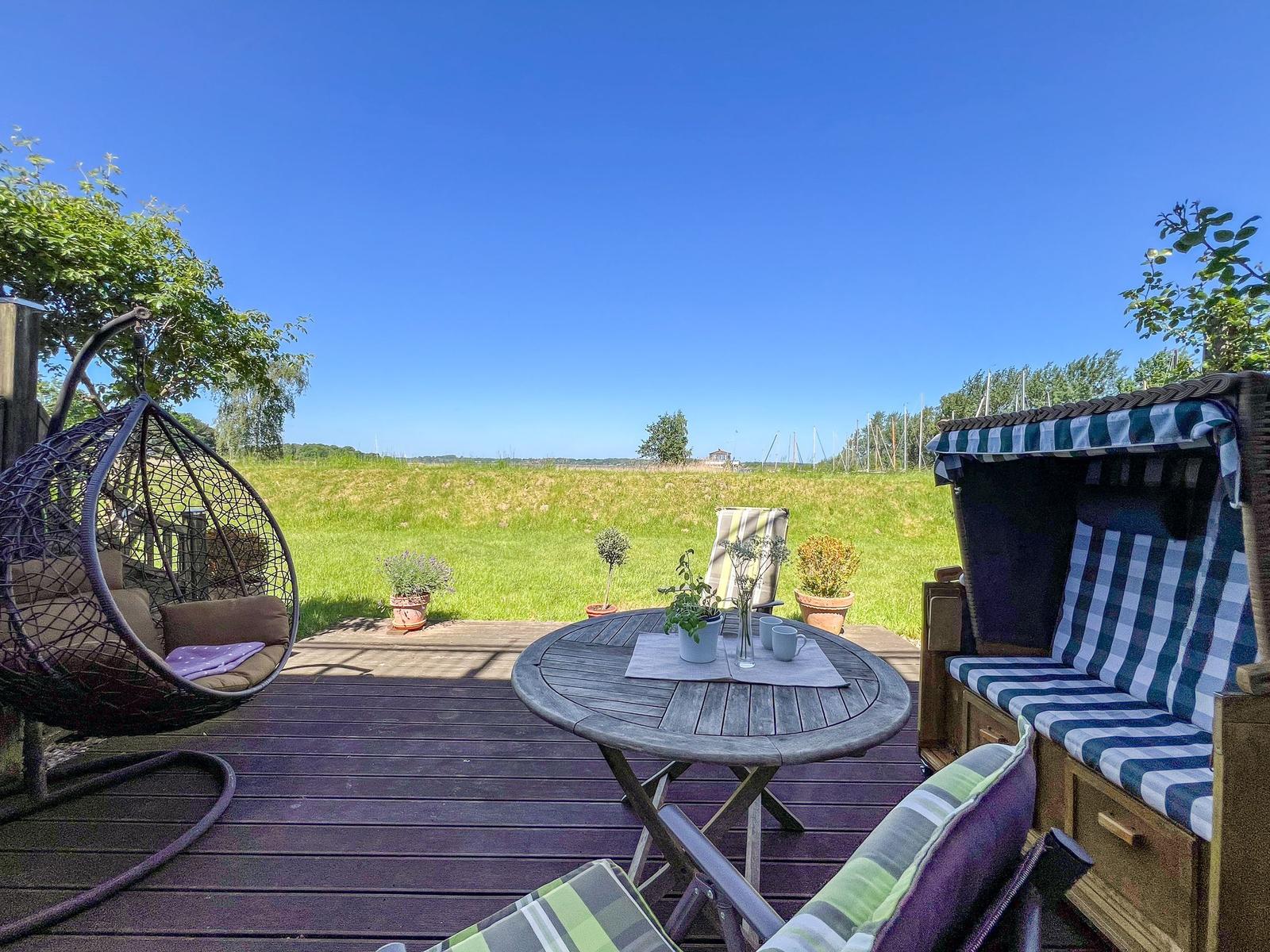 Terrasse mit Hängesessel, Tisch und Strandkorb. Blick auf grünes Feld unter blauem Himmel.