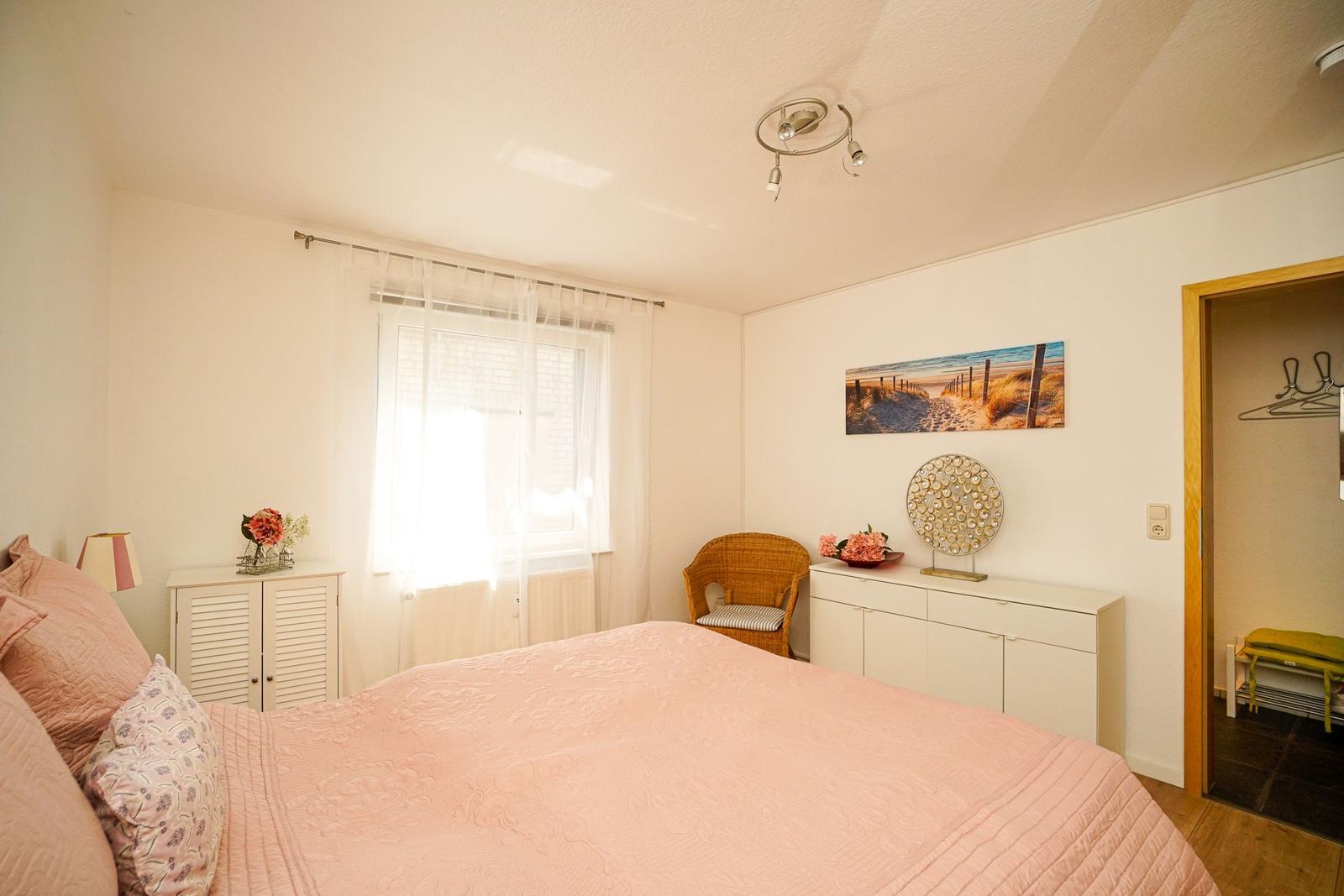 Schlafzimmer mit Bett, Schrank und Stuhl. Fenster mit Vorhängen und Wandbild.