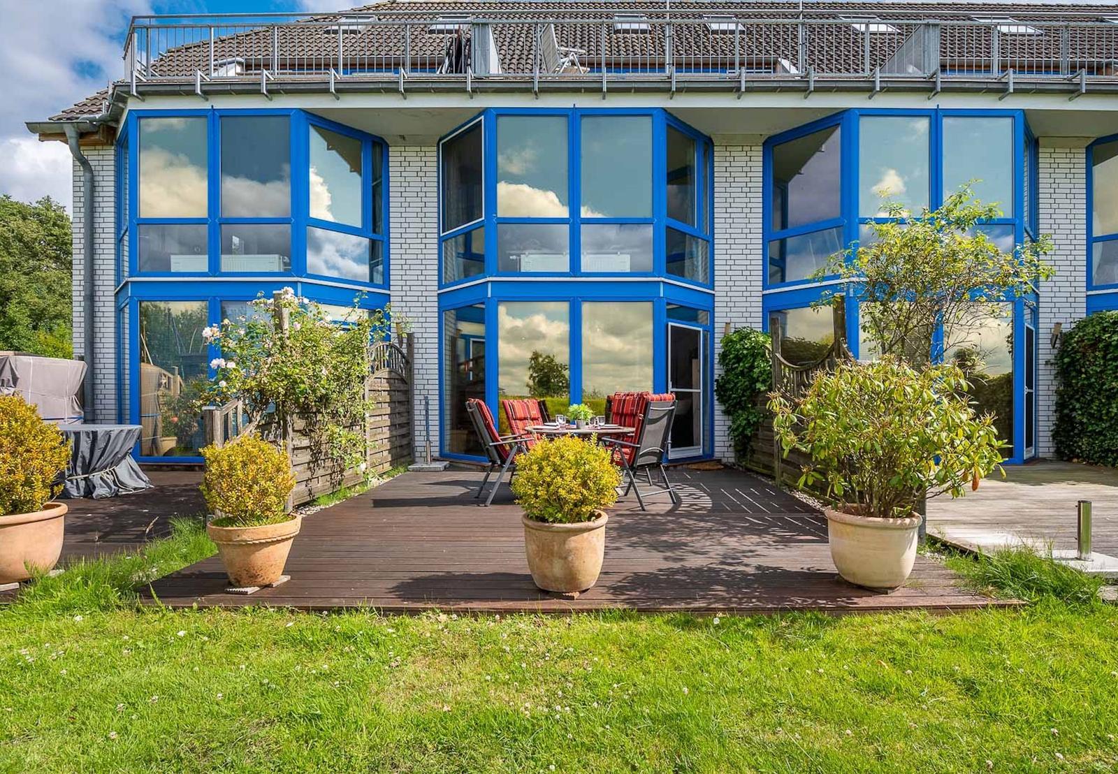 Haus mit blauen Fenstern und Terrasse mit Gartenmöbeln und Pflanzen.