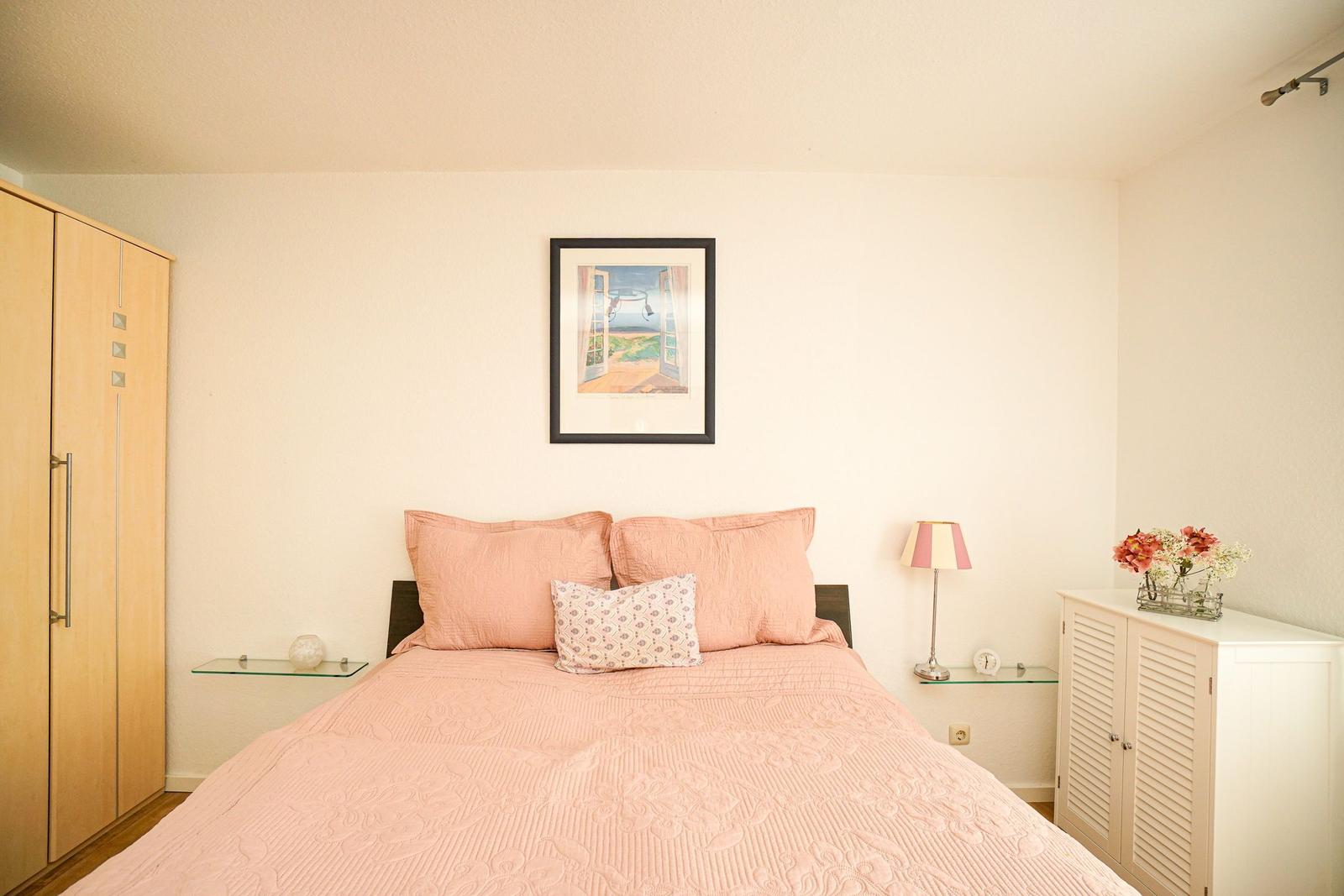 Schlafzimmer mit Bett, Kleiderschrank und Nachttisch. Wandbild und Blumen dekorieren den Raum.