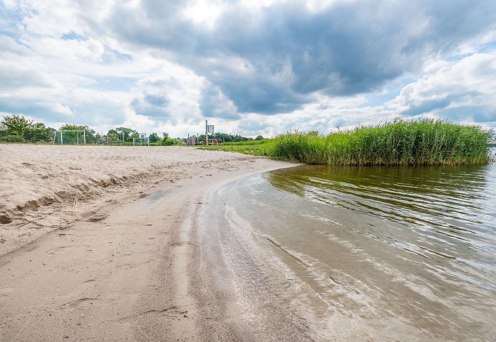 Strand mit Sand, Wasser und Gras. Im Hintergrund sind Bäume und ein Fußballtor zu sehen.