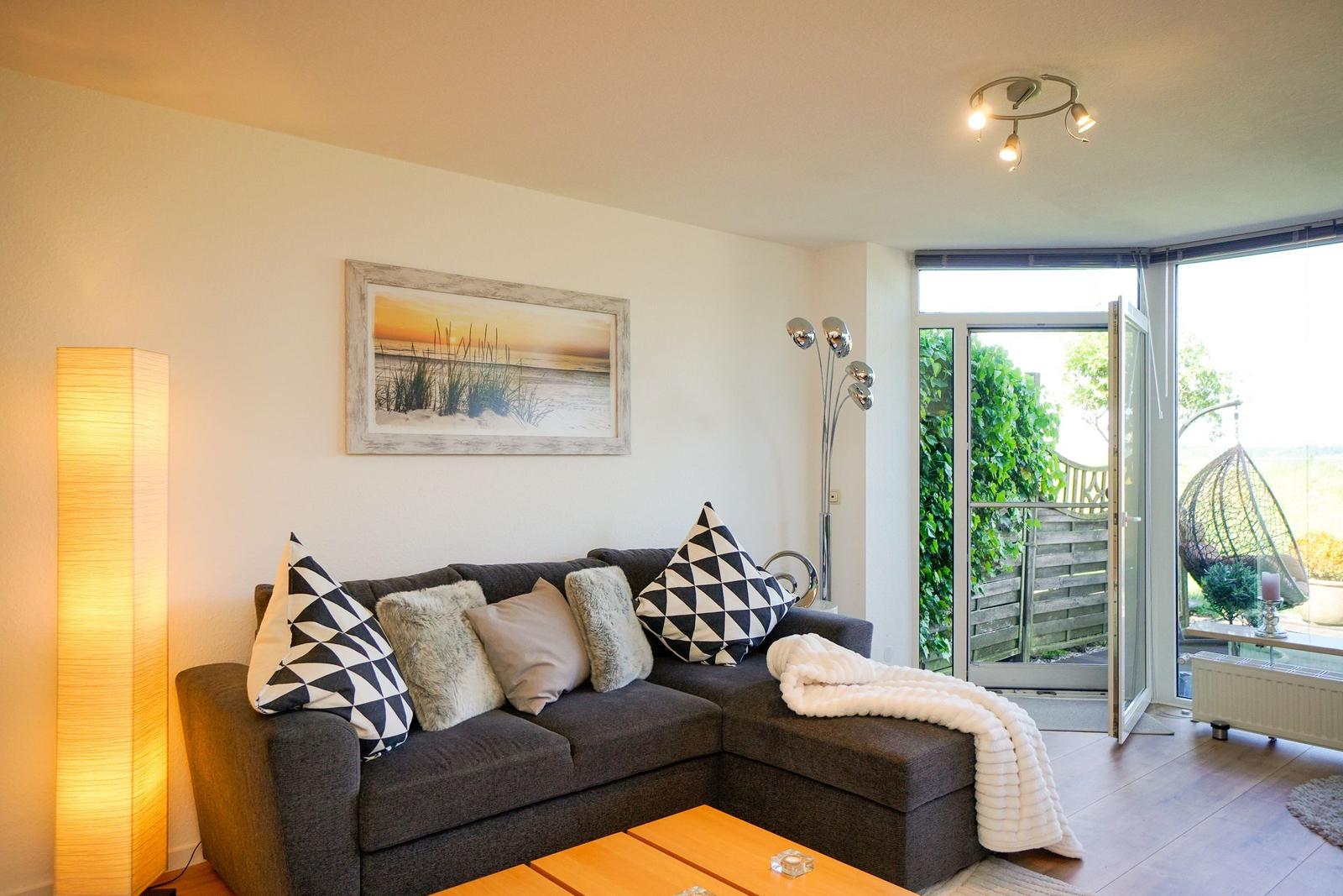 Ein Wohnzimmer mit einem grauen Sofa, Kissen und einer weißen Decke. Eine Glastür führt auf eine Terrasse.
