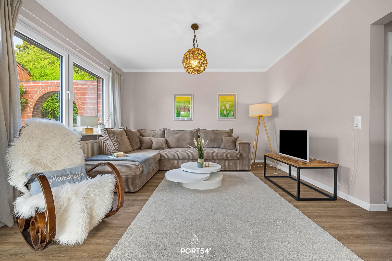 Wohnzimmer mit Sofa, Fernseher und Sessel. Fenster mit Blick auf Garten.
