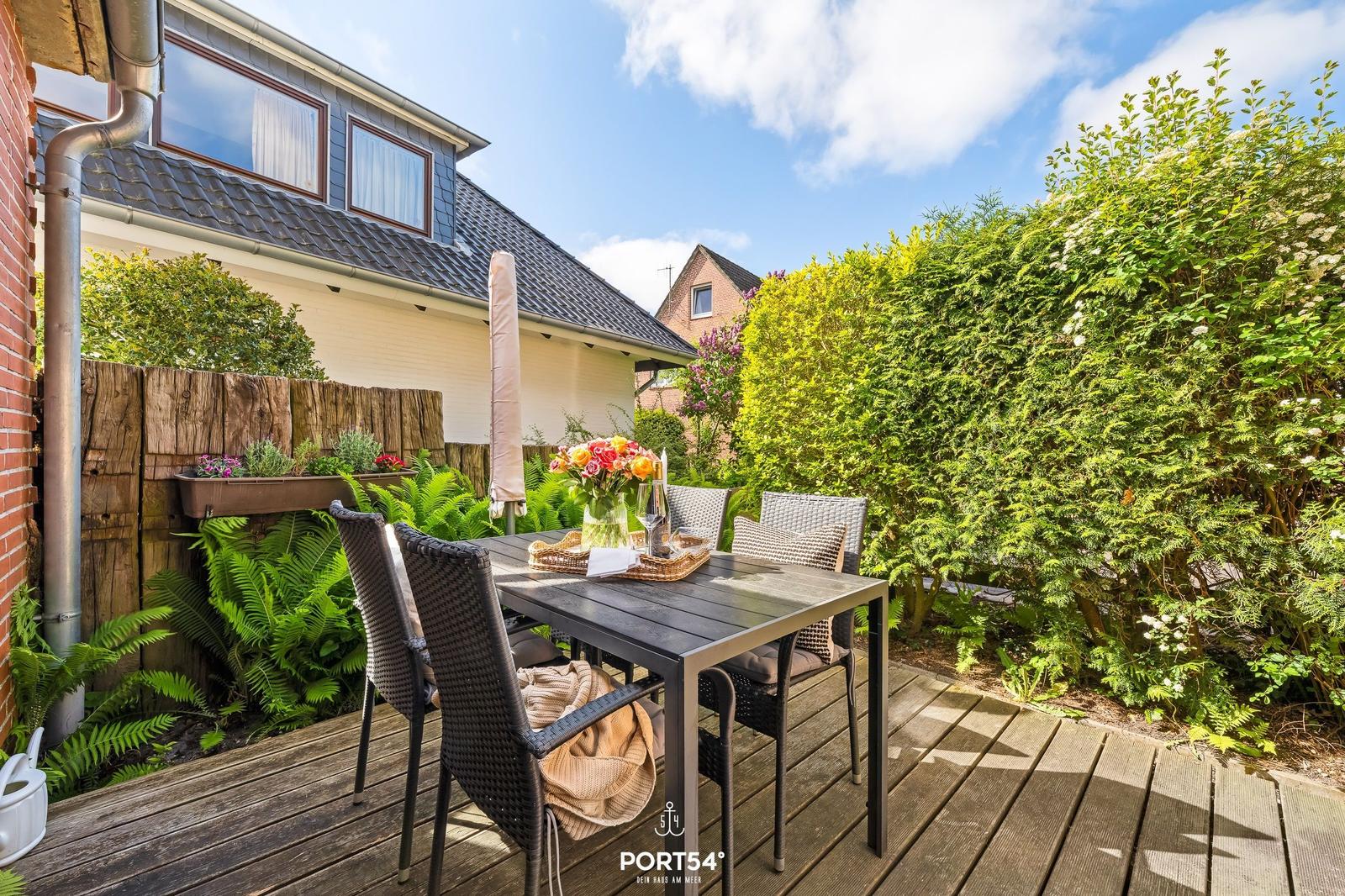 Terrasse mit Tisch, Stühlen und Blumenarrangement. Umgeben von Grün und Holzhauswand.