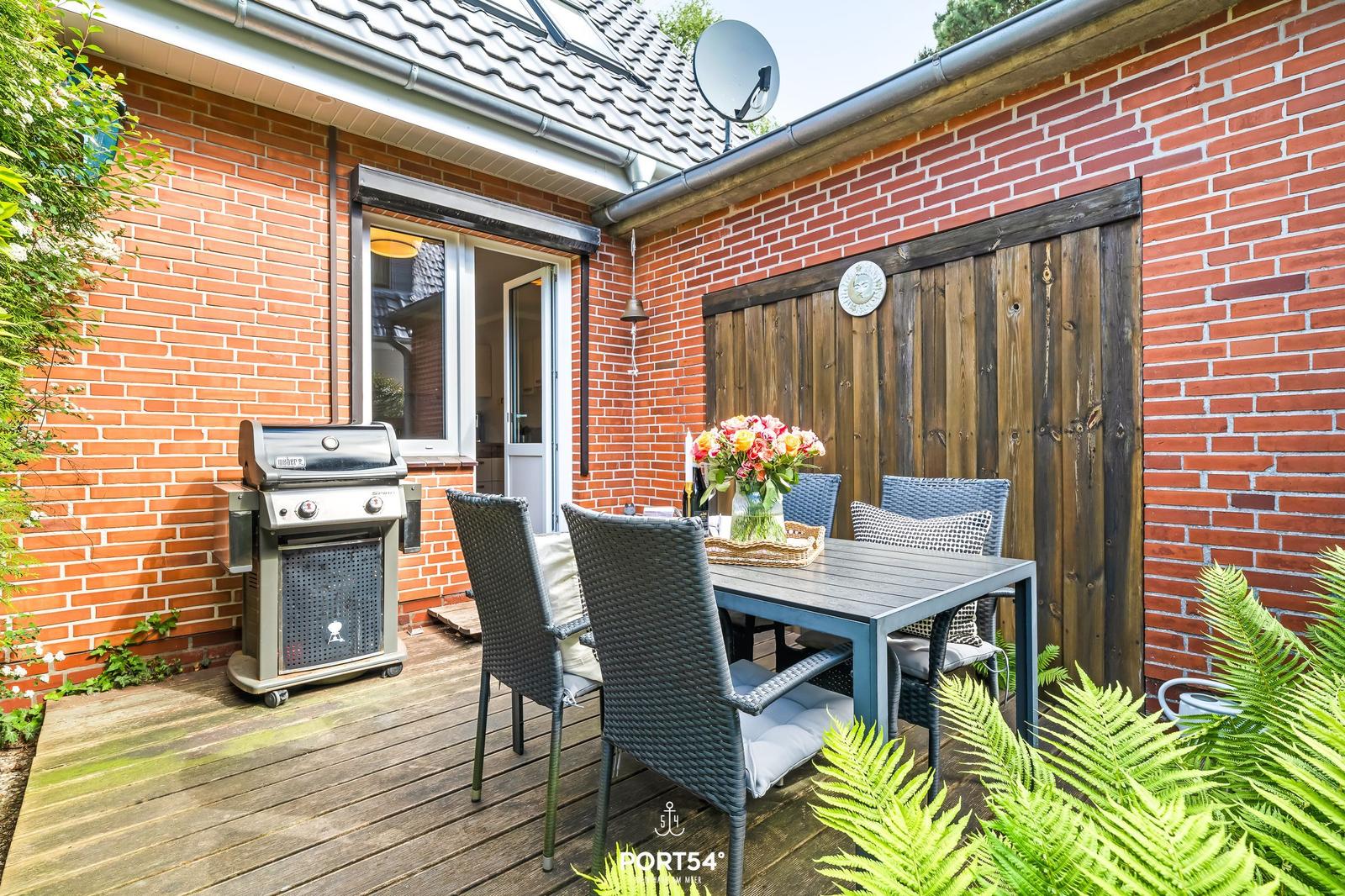 Terrasse mit Grill, Tisch und Stühlen. Roter Backsteinbau mit Holztür.