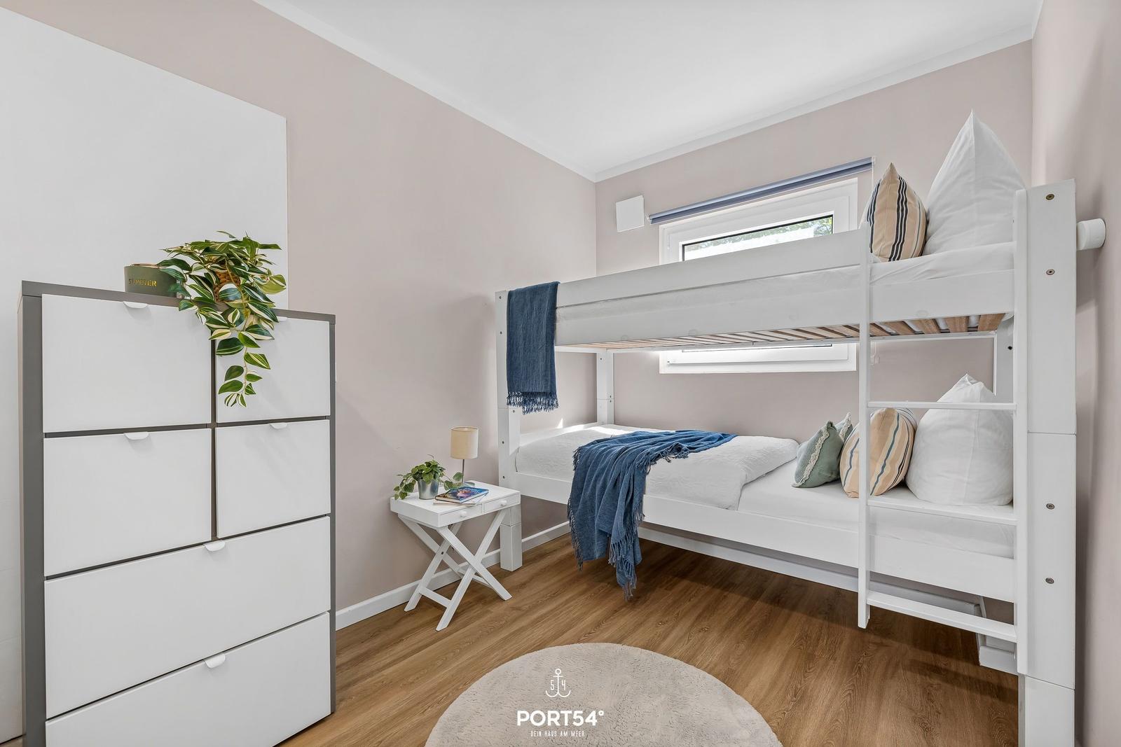 Schlafzimmer mit Doppelstockbett, Schrank und Nachttisch. Holzboden und Fenster sichtbar.