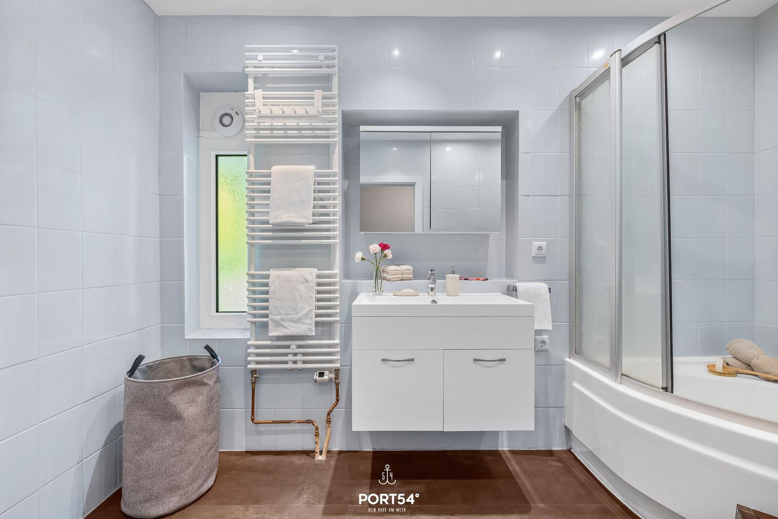 Badezimmer mit Dusche, Waschbecken und Heizung. Fenster und Handtuchhalter sichtbar.