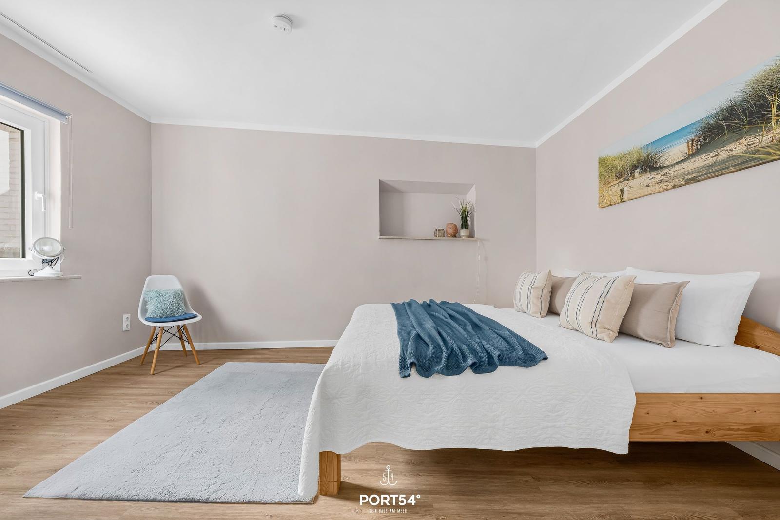 Schlafzimmer mit Bett, Stuhl und Wandbild. Holzboden und Fenster.