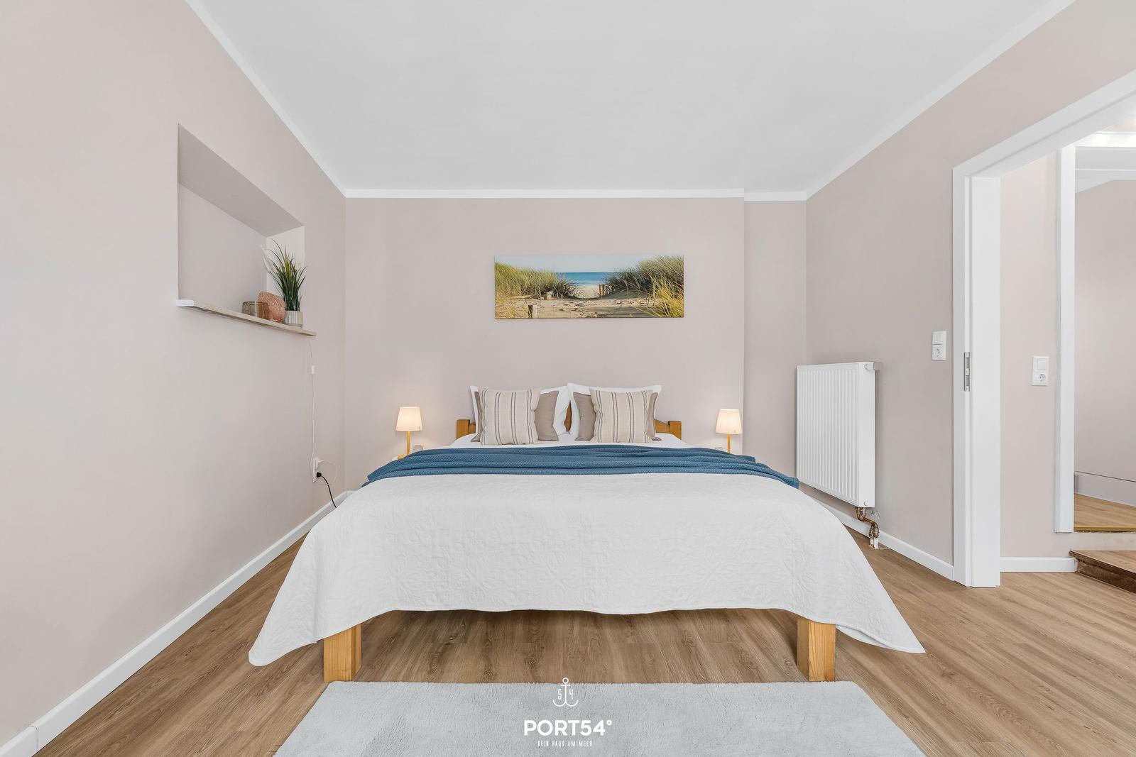 Schlafzimmer mit Bett, Wandbild und Holzboden. Tür führt in einen anderen Raum.