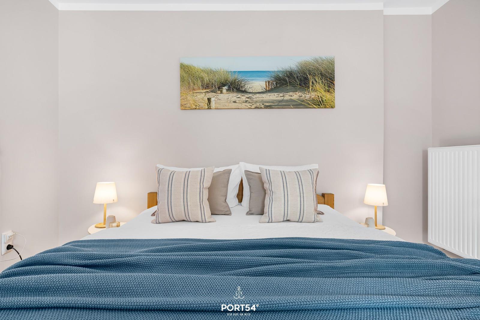 Schlafzimmer mit Bett, Kissen und Wandbild. Zwei Nachttische mit Lampen.