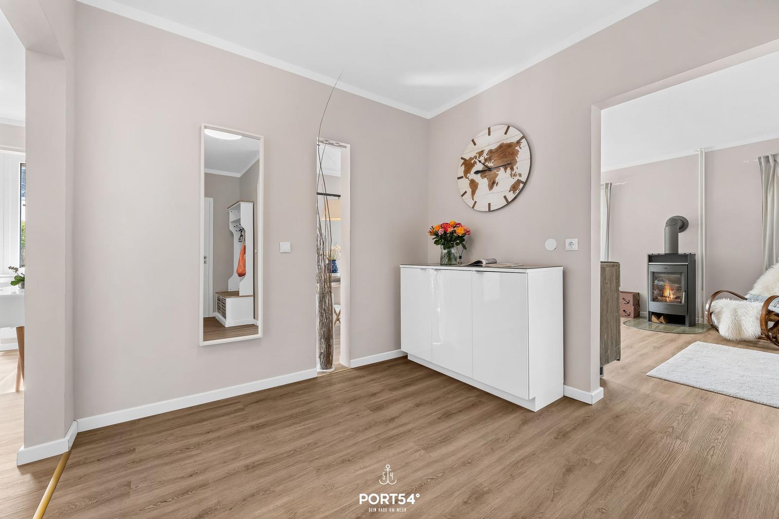 Eingangsbereich mit weißen Schränken, Wanduhr und Spiegeln. Blick in Wohnzimmer mit Kamin.
