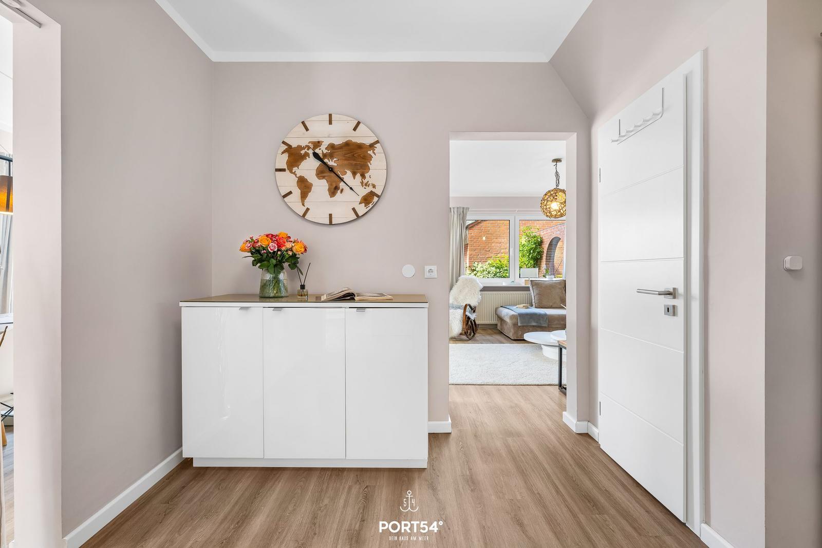 Eingangsbereich mit weißen Schränken, Weltuhr und Blumen. Durchgang zum Wohnzimmer mit Sofa.