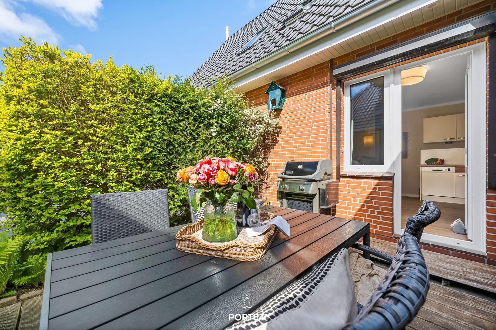 Terrasse mit Tisch, Stühlen und Grill. Blick in die Küche. Grüner Heckenzaun.