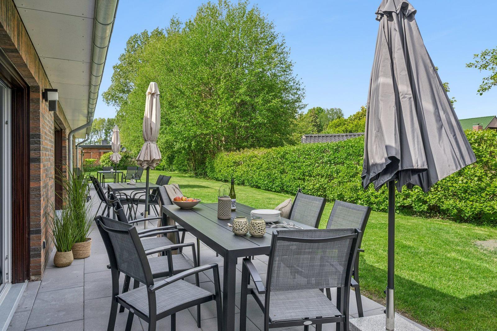 Terrasse mit Tisch, Stühlen und Sonnenschirmen. Grünflächen und Bäume im Hintergrund.
