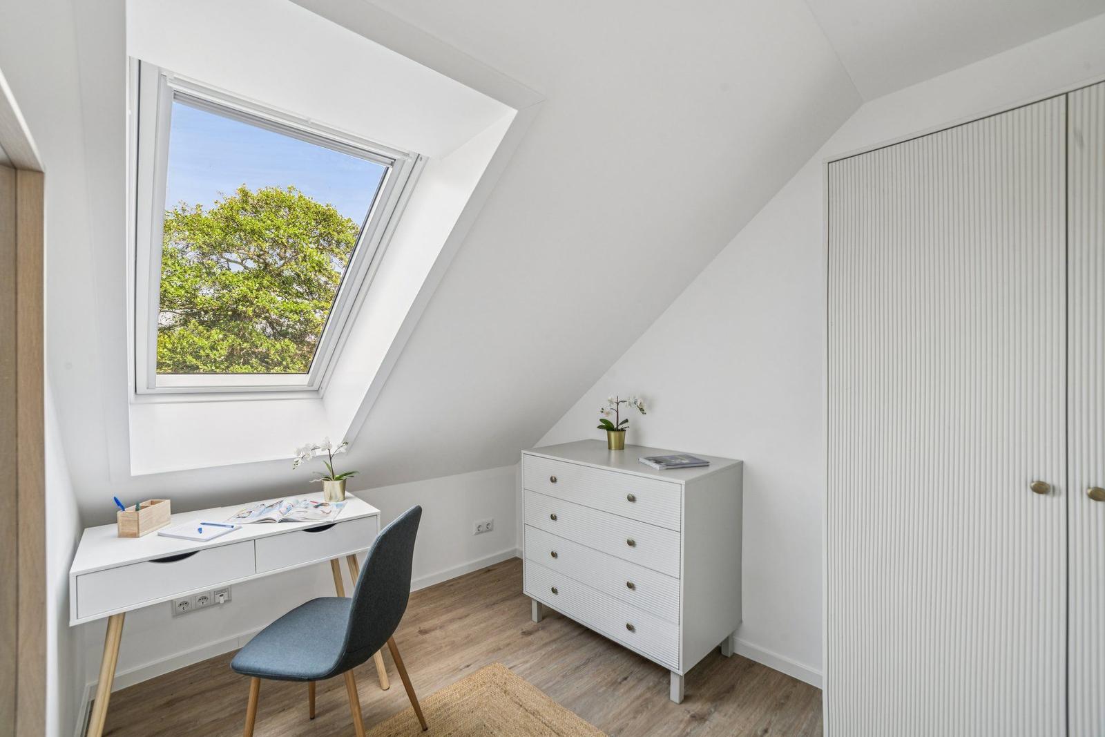 Dachgeschosszimmer mit Schreibtisch, Kleiderschrank und Dachfenster mit Blick auf Bäume.