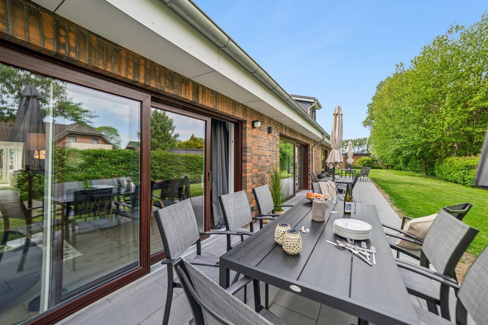 Terrasse mit Tisch, Stühlen und Sonnenschirmen neben grünem Garten.