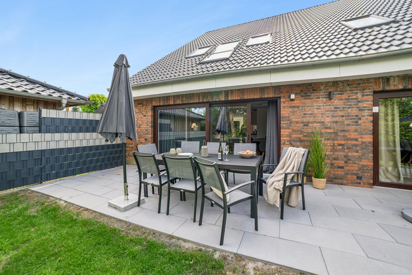 Terrasse mit Tisch, Stühlen und Schirm. Hintergrund: Haus mit Glasfront und Dachfenstern.