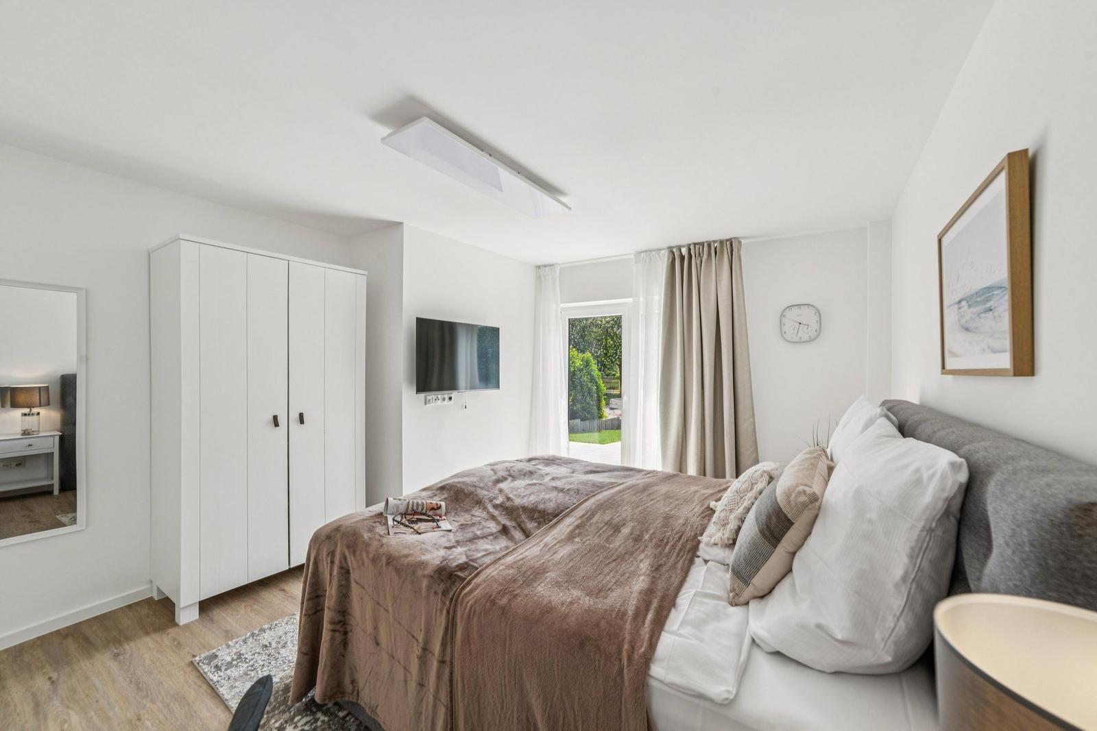 Schlafzimmer mit Bett, Kleiderschrank, Fernseher und Fenster mit Blick auf Garten.