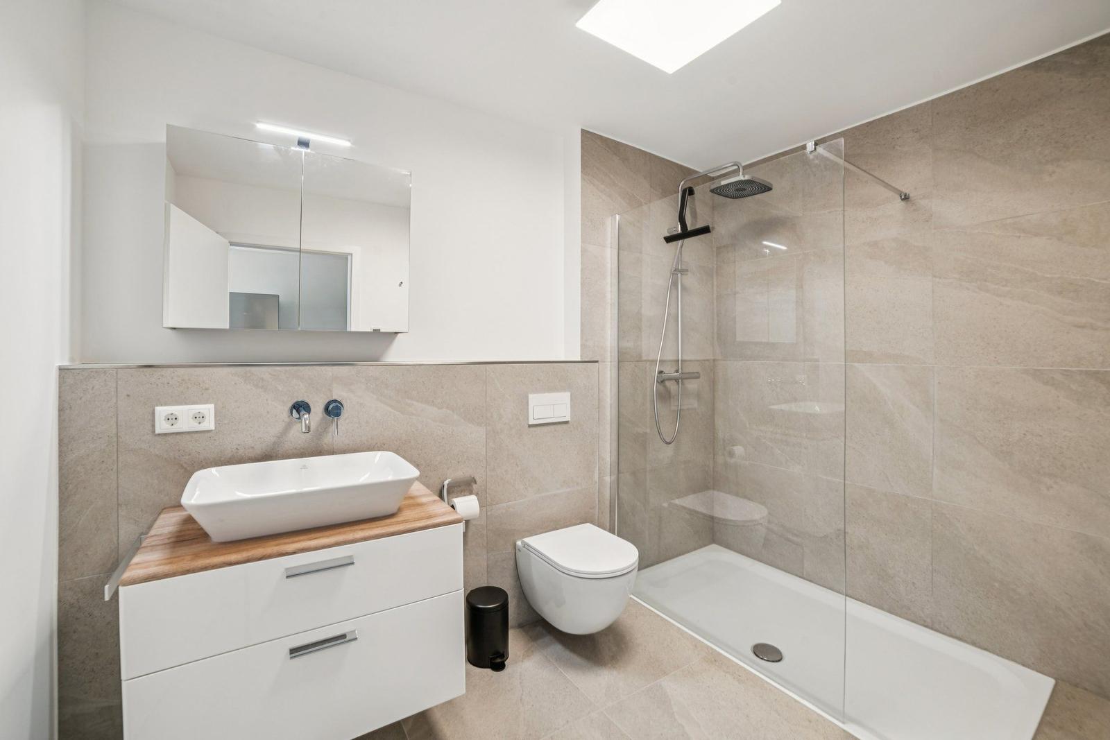 Badezimmer mit Dusche, Waschbecken und Toilette. Fliesenwand und Spiegelschrank.