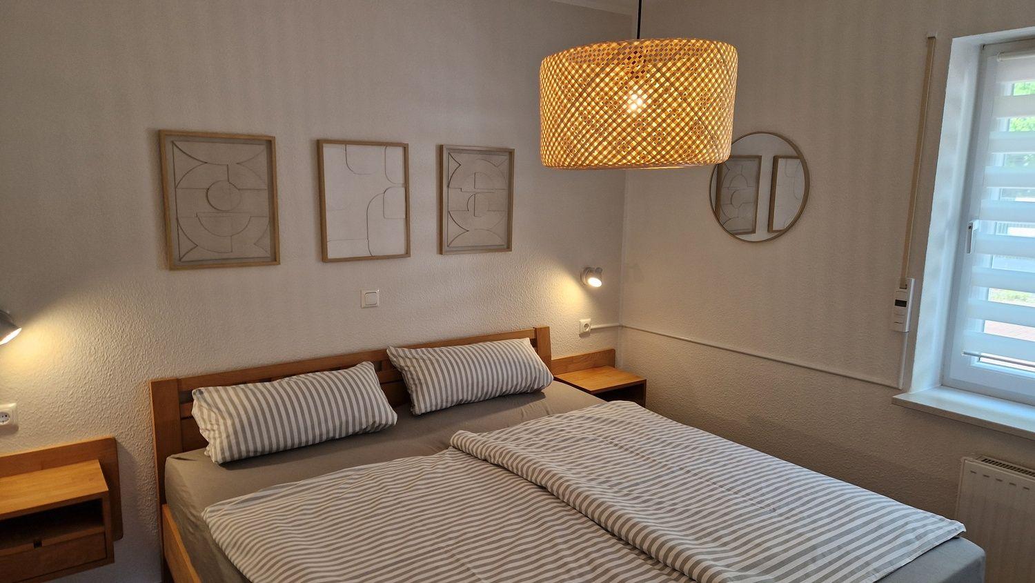 Doppelbett mit Streifenbezug, Wandbilder und Holz-Nachtisch. Rundspiegel und Hängelampe.