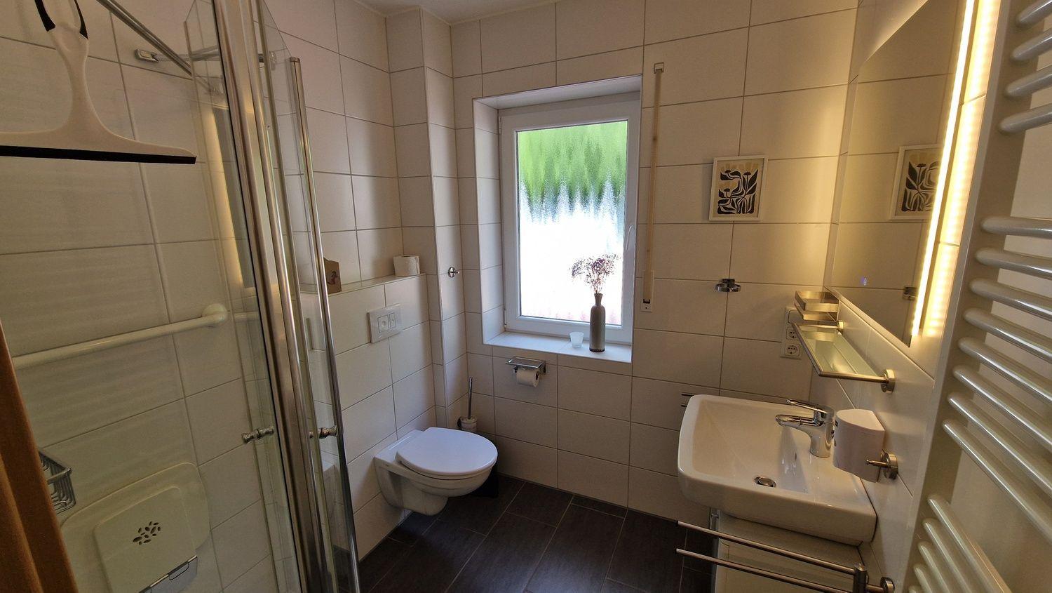 Badezimmer mit Dusche, Toilette und Waschbecken. Fenster mit Blumenvase.