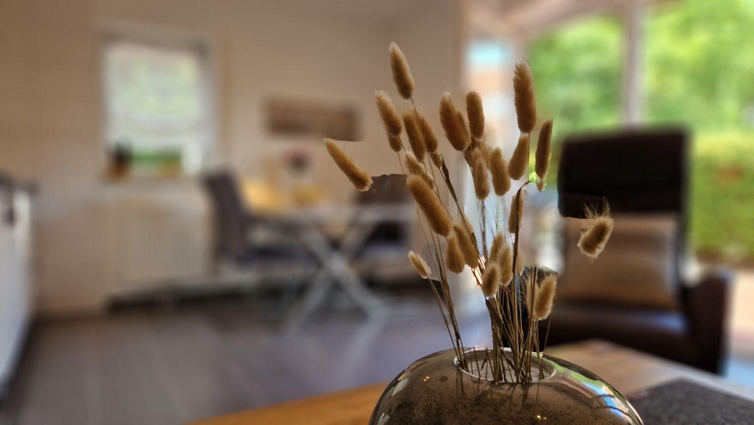 Trockenblumen in einem Glas auf einem Tisch. Im Hintergrund ist ein Wohnbereich mit Sitzmöbeln und Fenstern.