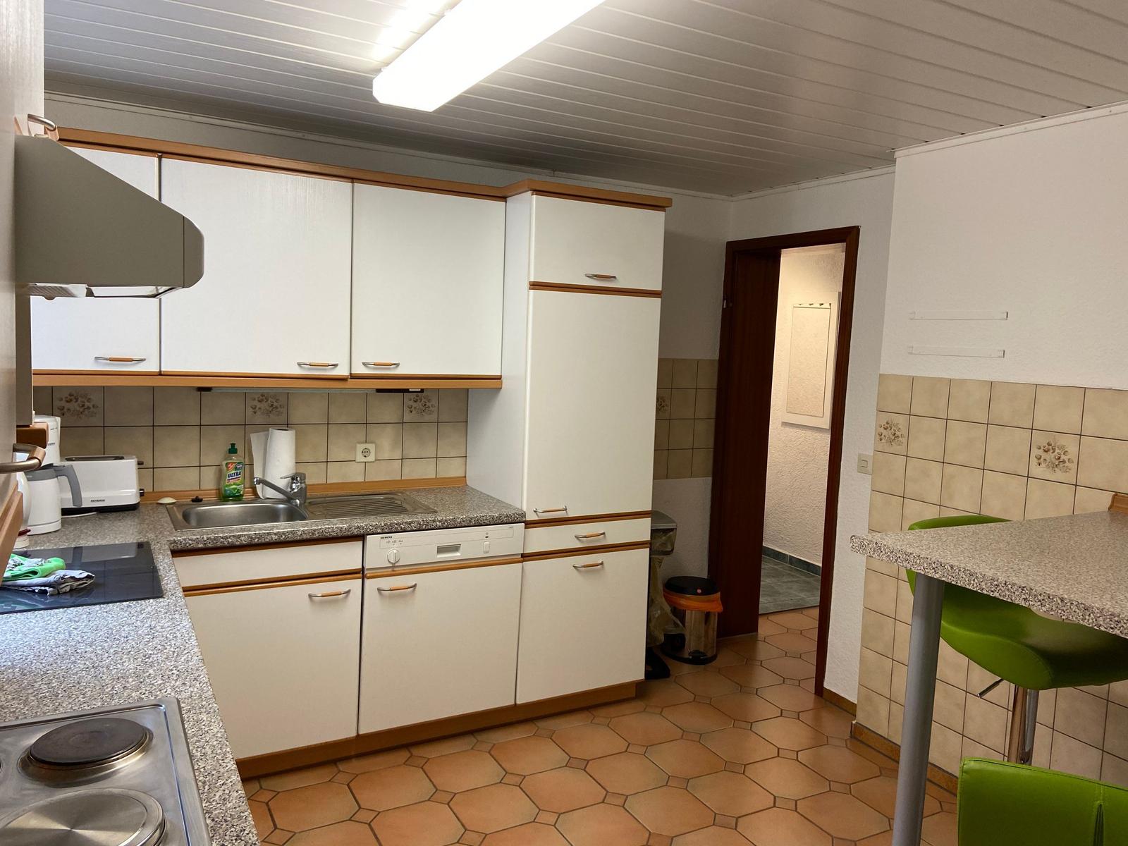 Küche mit weißen Schränken, Spüle und Arbeitsfläche. Grün sitzende Barhocker.