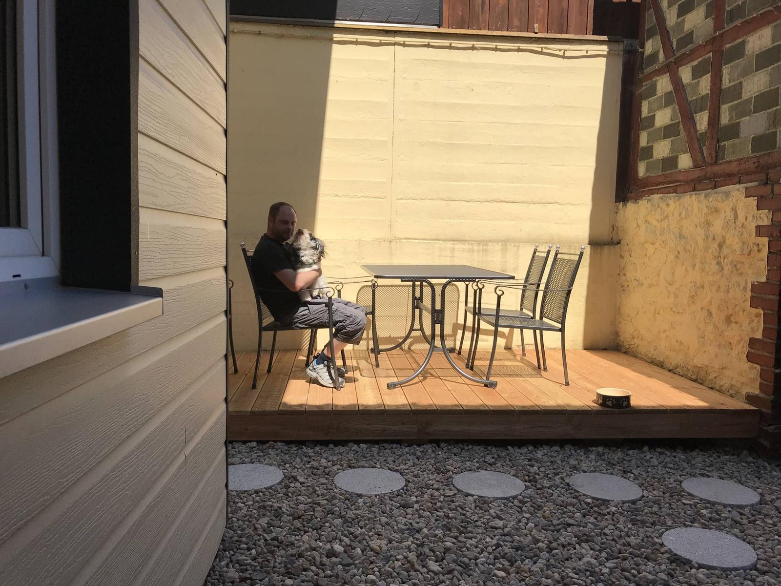 Mann mit Hund sitzt auf Terrasse mit Tisch und Stühlen.