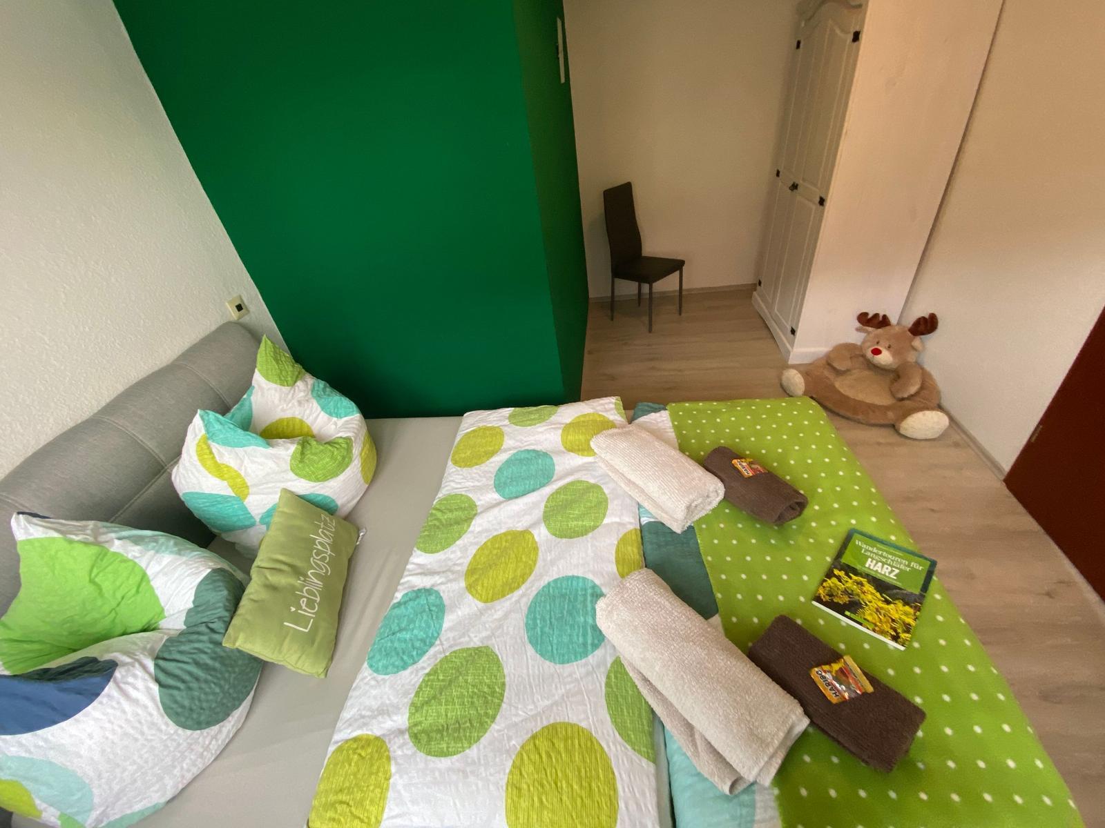 Schlafzimmer mit Bett, Kissen und Handtüchern. An der Wand ein grüner Bereich.