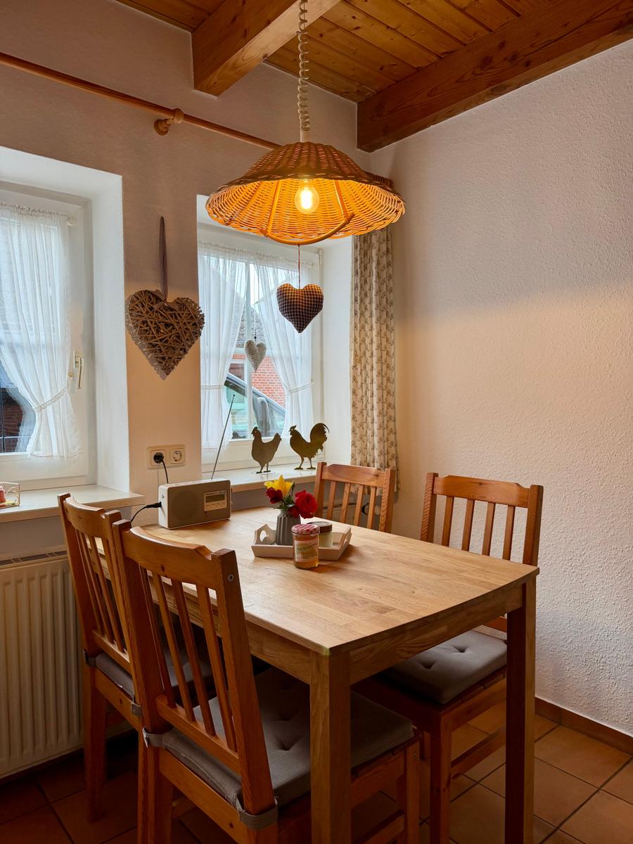 Holztisch mit vier Stühlen unter einer Wickerlampe. Fenster mit Vorhängen und Dekoration.
