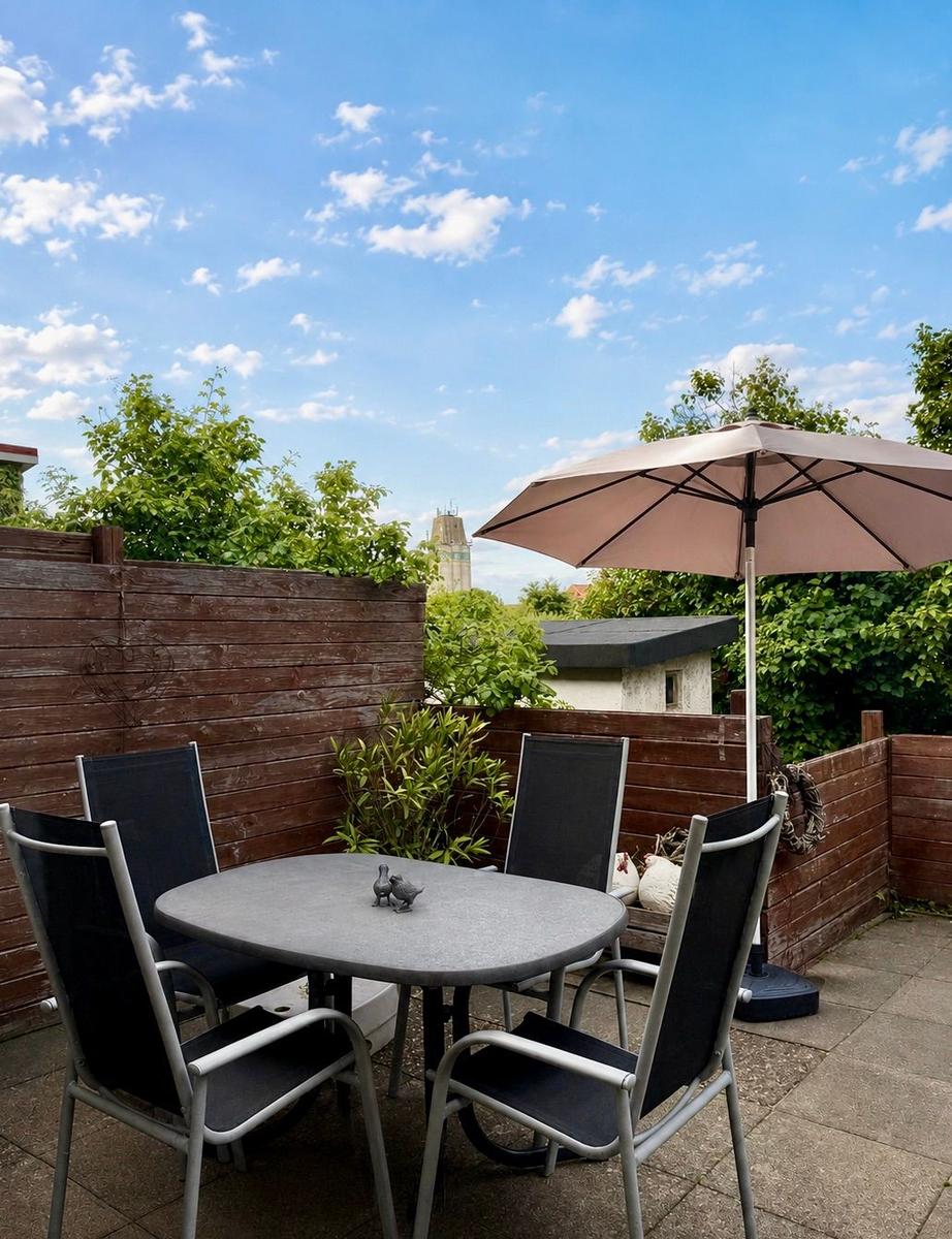 Terrasse mit Tisch, vier Stühlen und Sonnenschirm. Holzumrandung und grüne Pflanzen im Hintergrund.