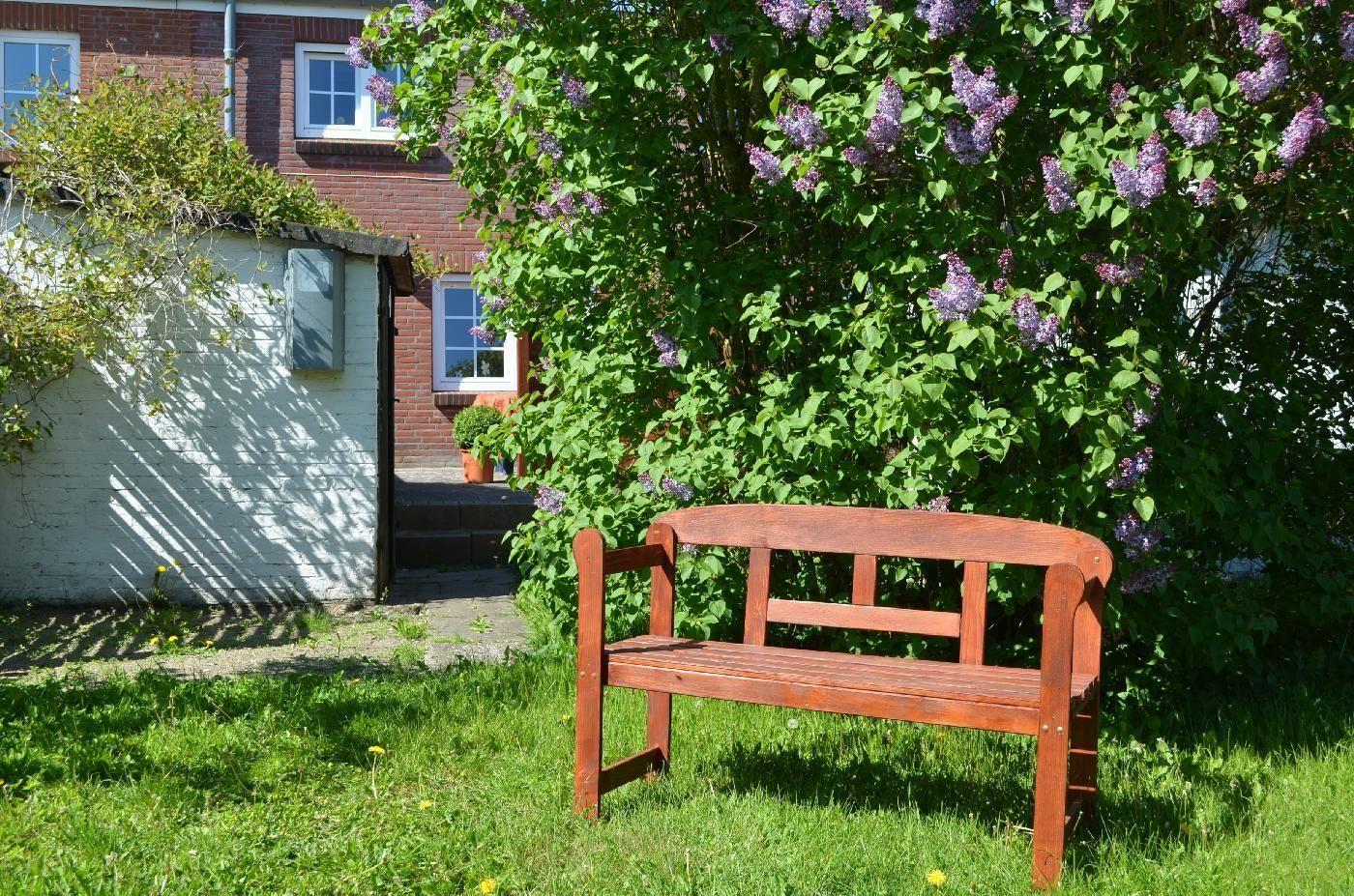Holzbank im Garten vor blühendem Lilienstrauch und rotem Backsteinhaus.