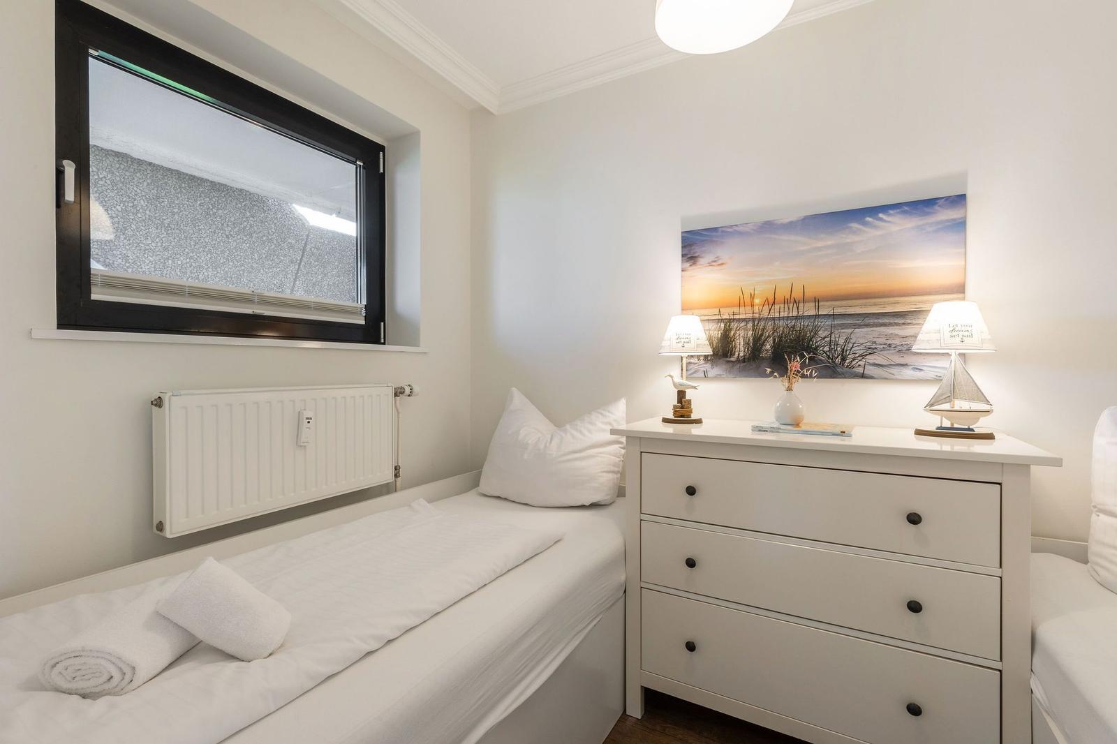 Ein Schlafzimmer mit Bett, Schrank und Fenster. Auf dem Schrank stehen zwei Lampen und ein Bild.