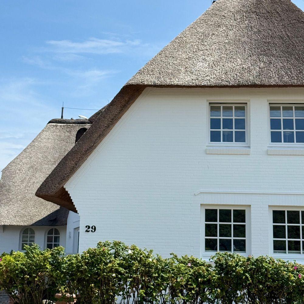 Weiße Hauswand mit Strohdach und Fenstern. Hausnummer 29. Grünbewachsener Vorgarten.
