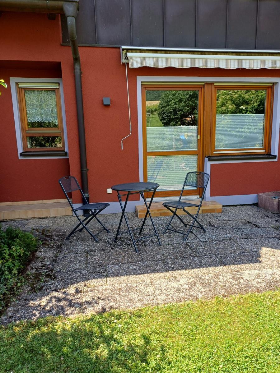 Außenbereich mit Tisch und zwei Stühlen vor rotem Haus mit Fenstern.