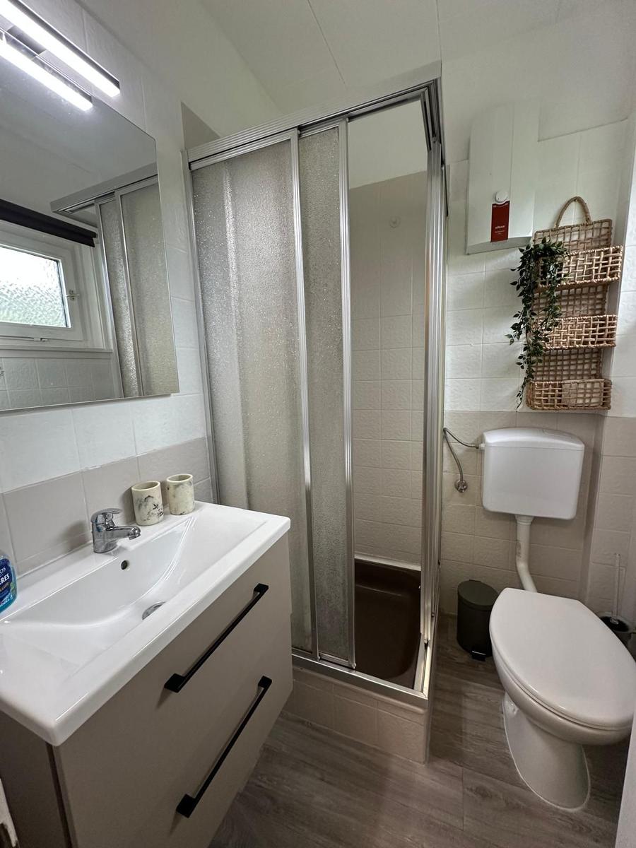 Badezimmer mit Dusche, Waschbecken und Toilette. Wandregal und Spiegel sichtbar.