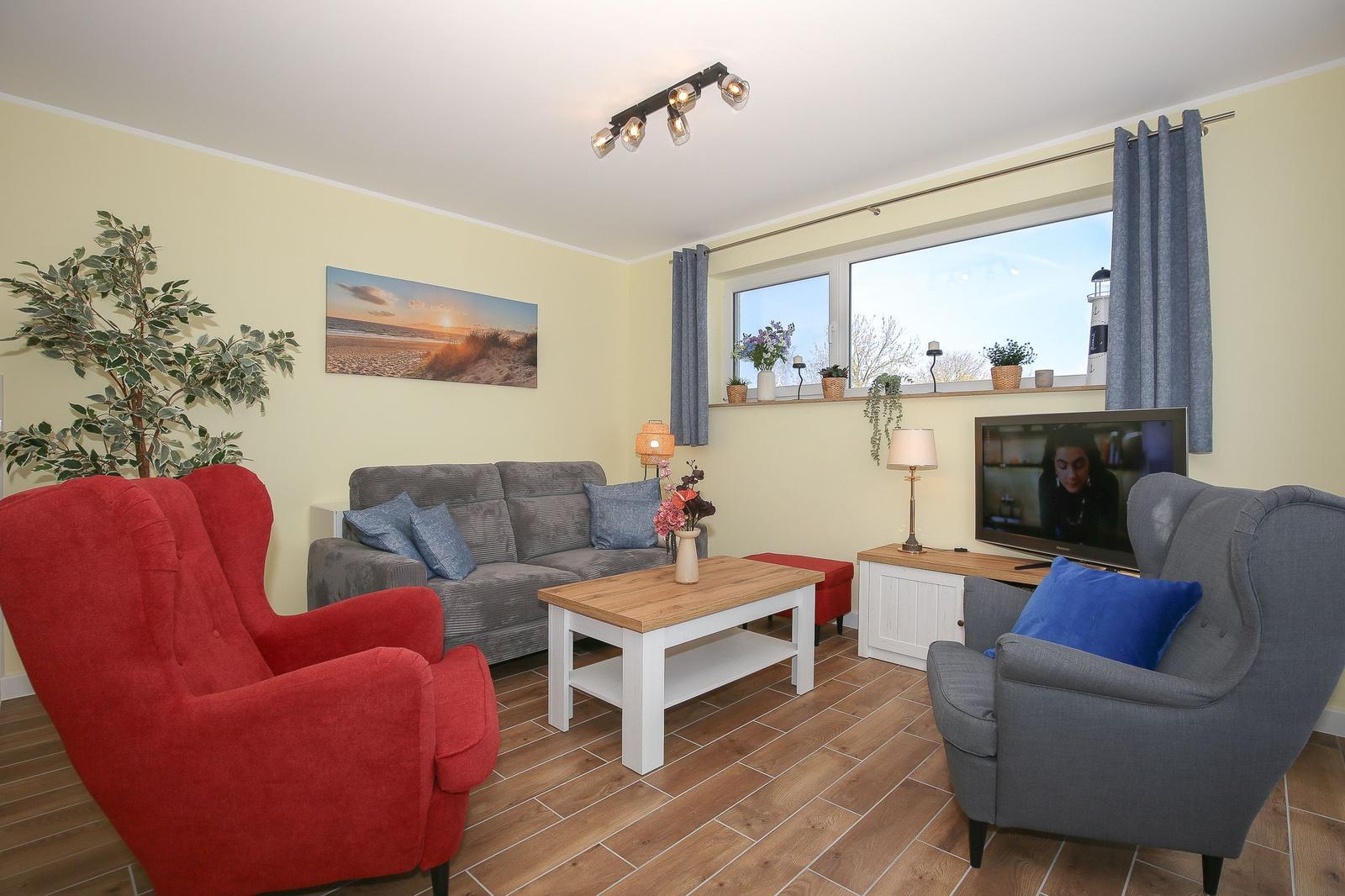 Wohnzimmer mit Sofa, zwei Sesseln, Couchtisch und TV. Fenster zeigt Leuchtturm.