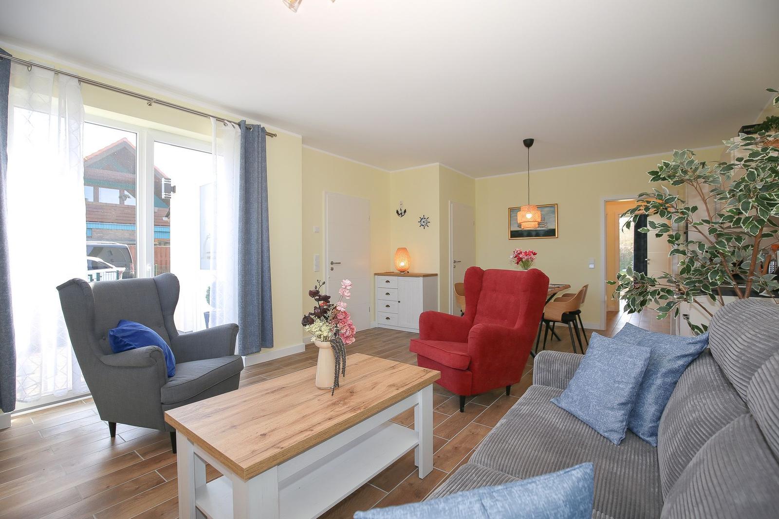 Wohnzimmer mit Sofa, Sessel und Esstisch. Große Fenster und Pflanzen.