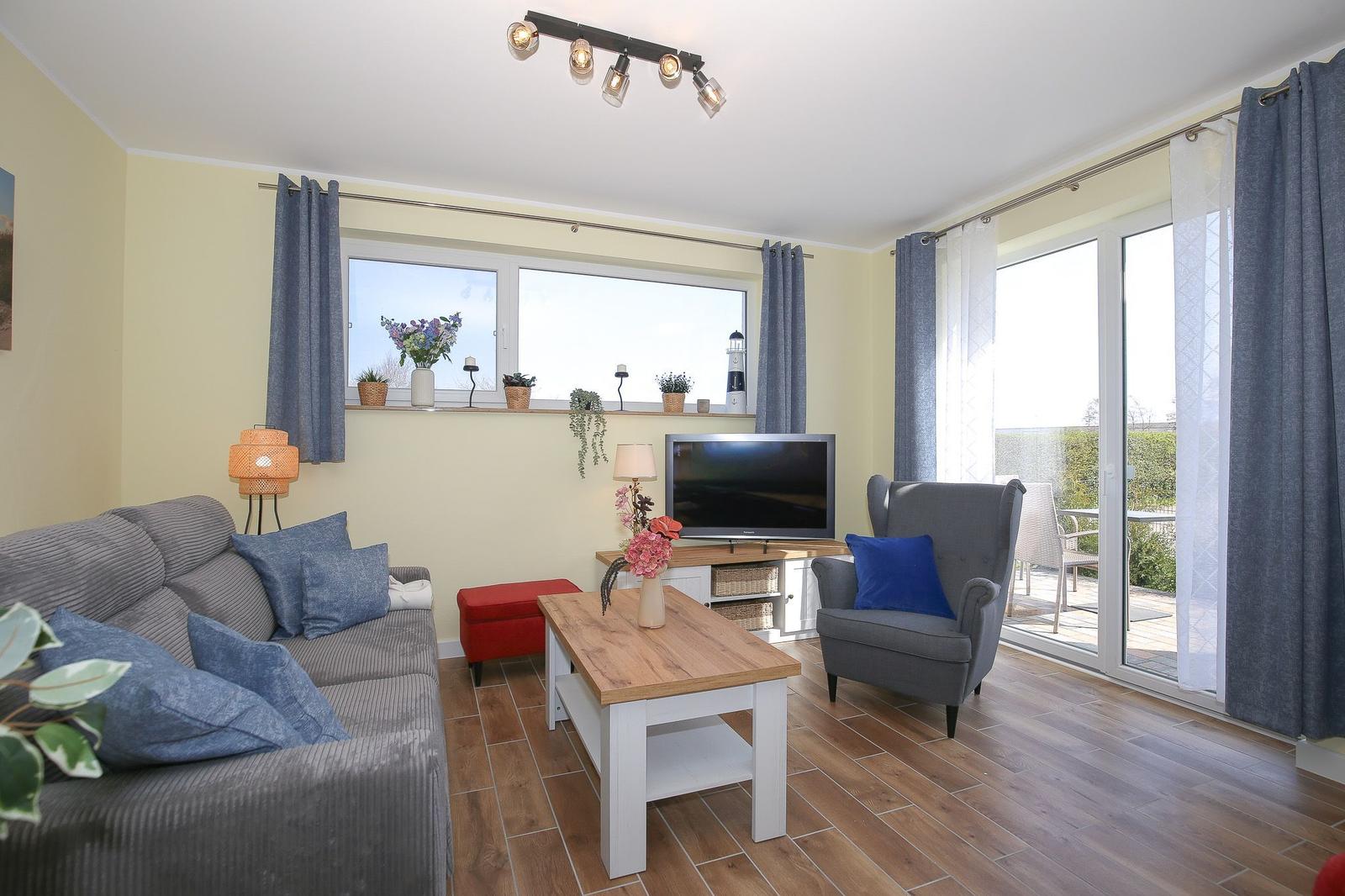 Wohnzimmer mit Sofa, Sessel und Fernseher. Fenster und Glastür führen auf eine Terrasse.