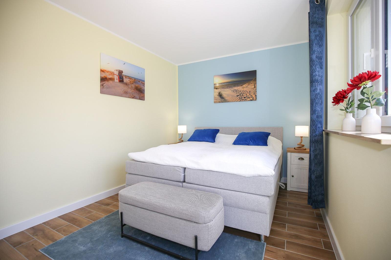 Schlafzimmer mit Bett, Nachttisch und Wandbildern. Fenster mit Blumen.