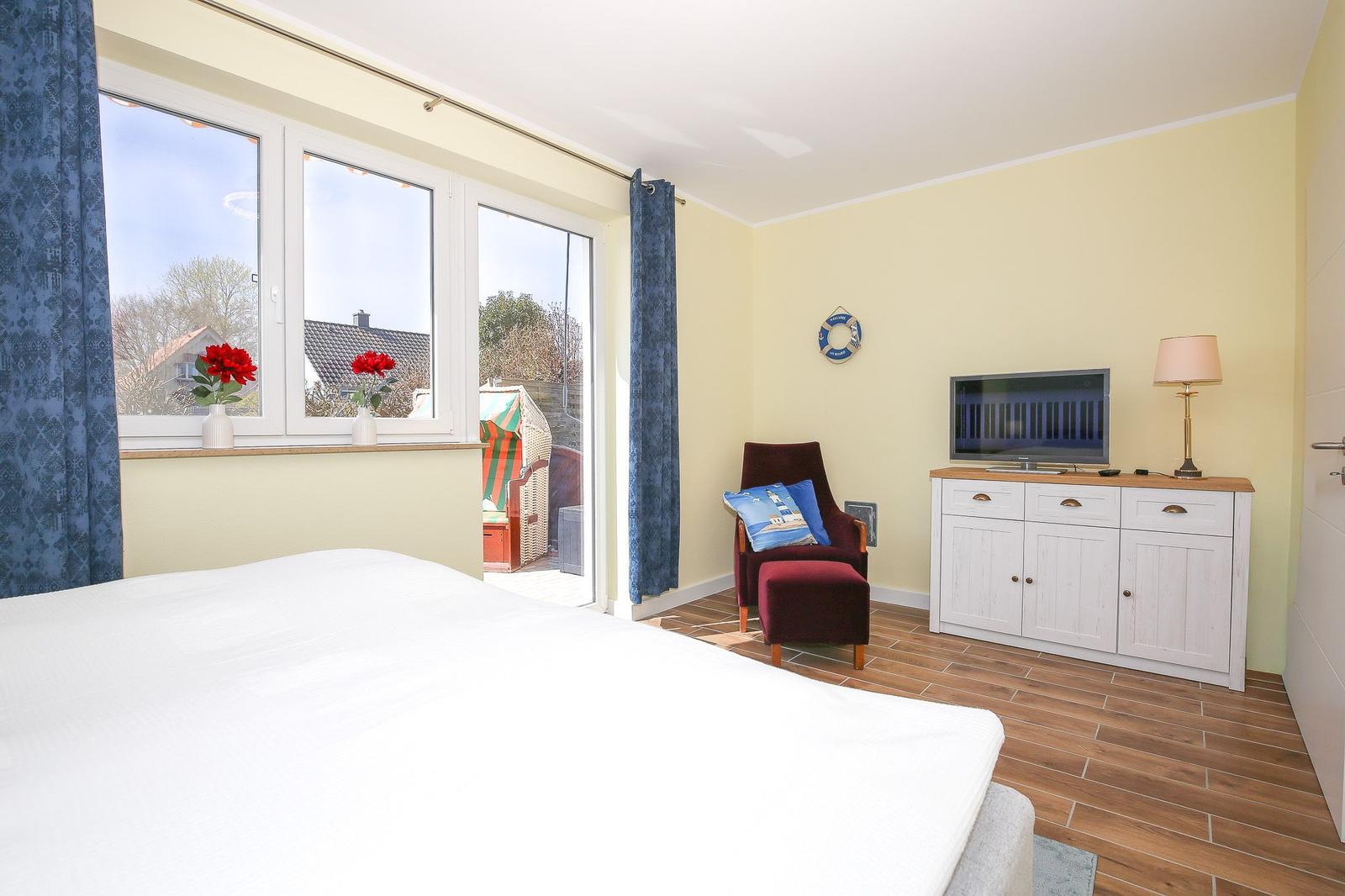 Schlafzimmer mit Bett, TV-Schrank, Sessel und Fenster zur Terrasse.