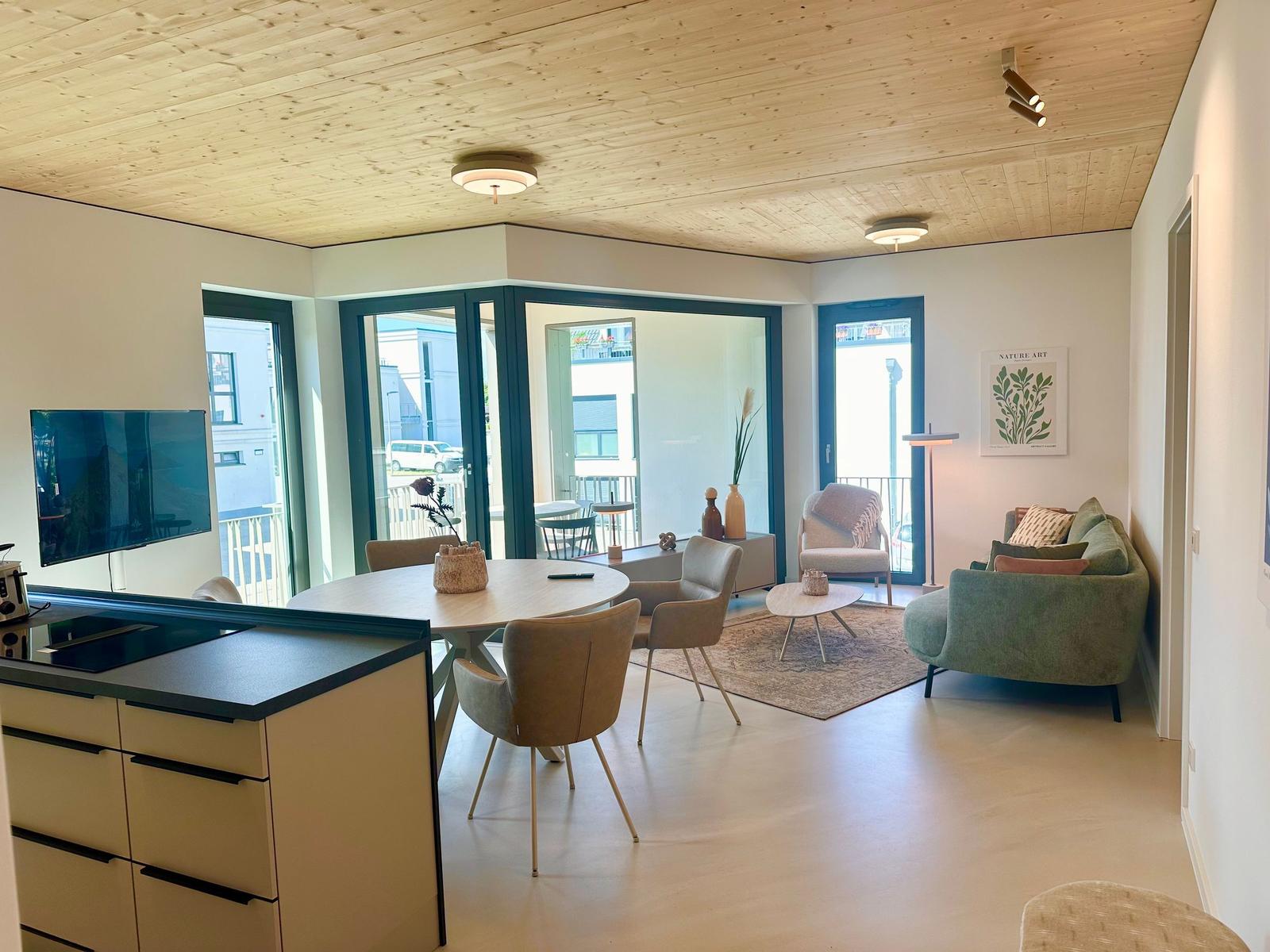 Wohn- und Essbereich mit Holzdecke, Sofa, Tisch und Fenstern zum Balkon.