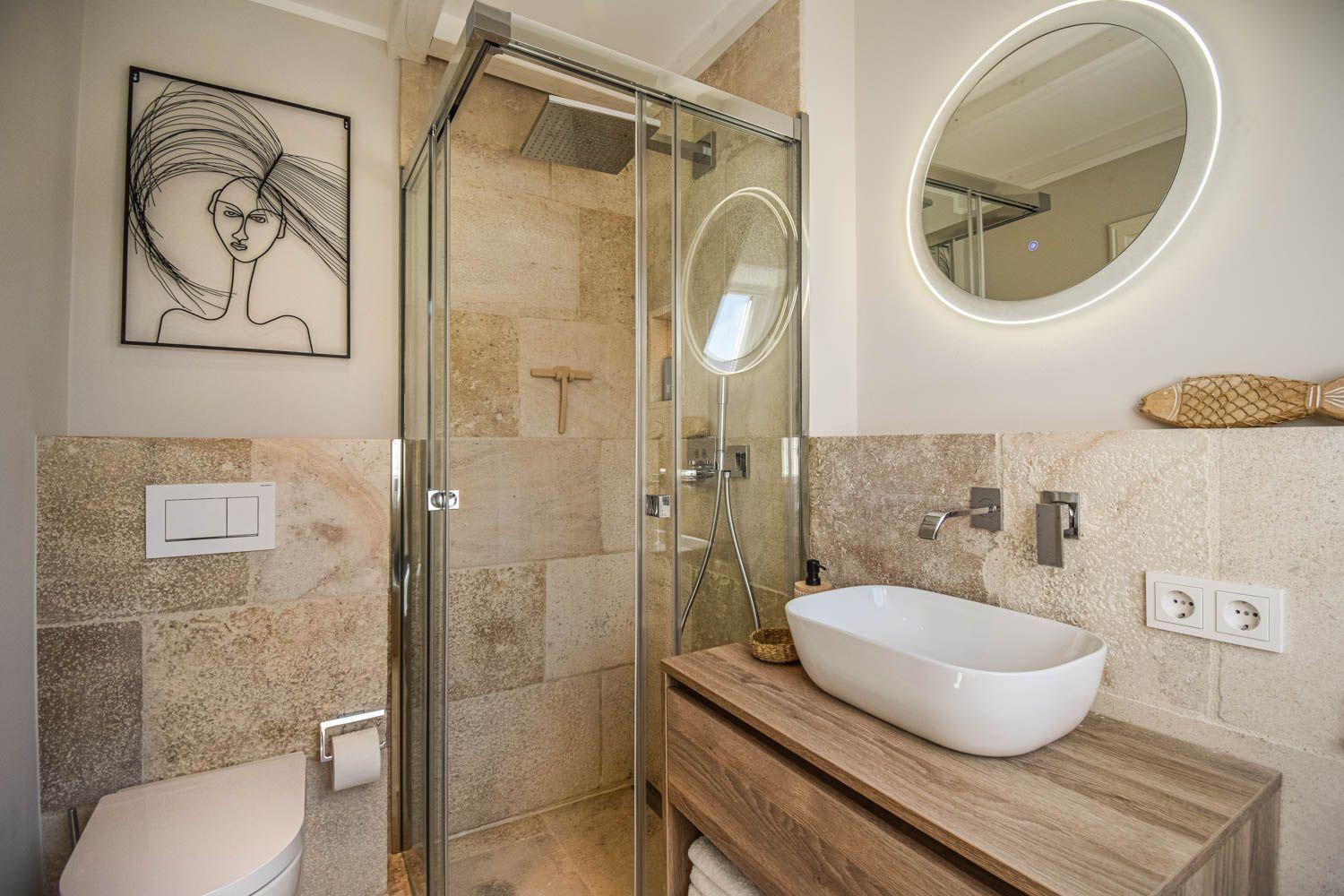 Badezimmer mit Dusche, Waschbecken und Toilette. Steinwand und runder Spiegel.