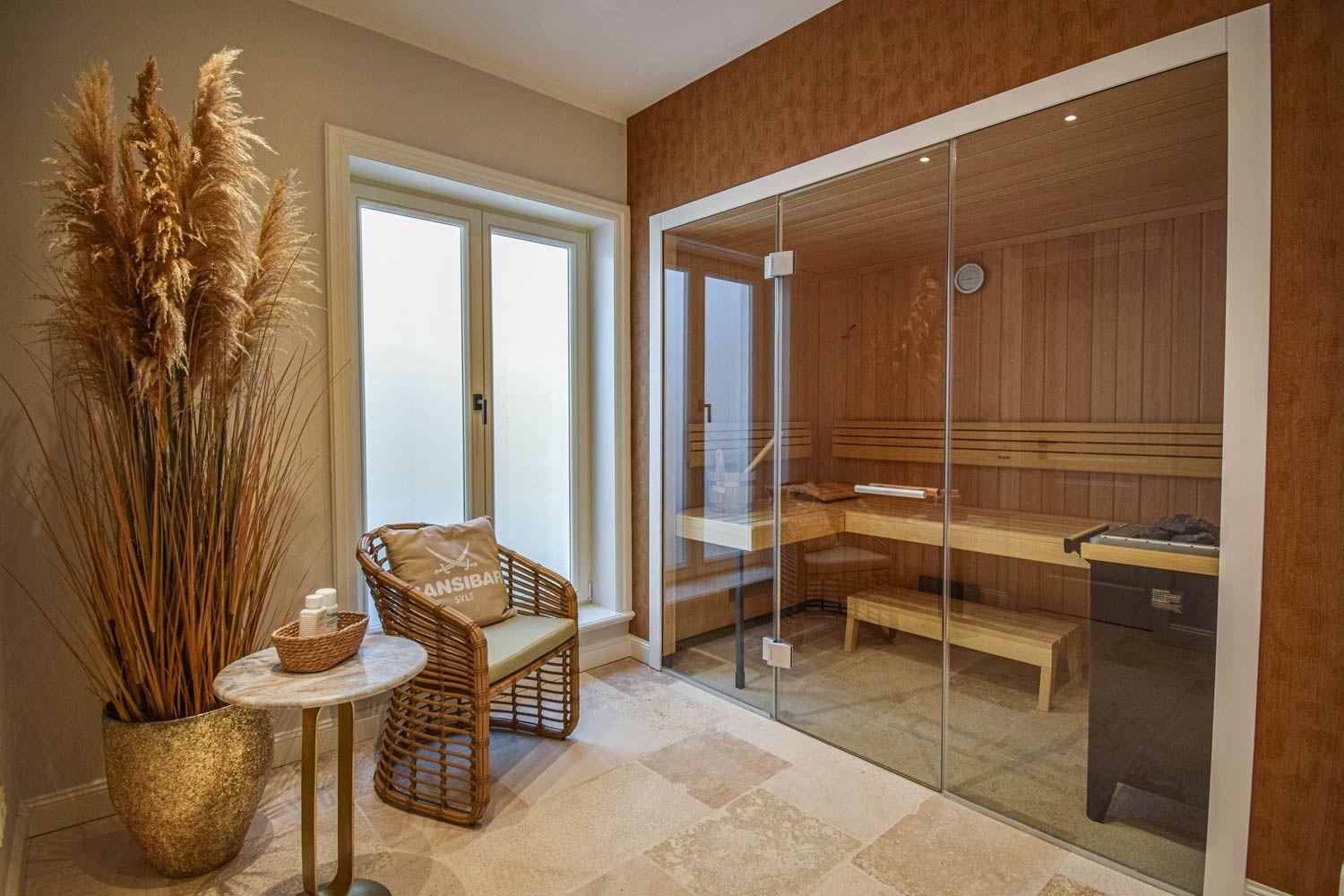 Sauna mit Glaswand und Holzbank. Nebenraum mit Sitzgelegenheit und Pflanze.