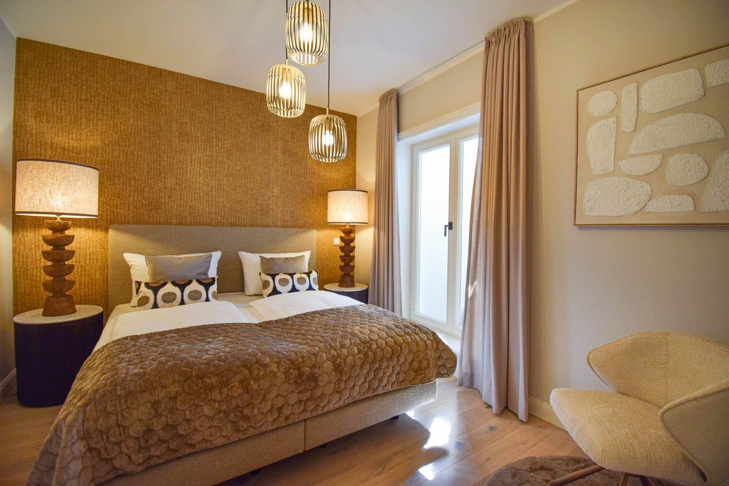 Schlafzimmer mit Bett, zwei Nachttischen, Lampe und Sessel. Wandverkleidung und Fenster mit Vorhängen.