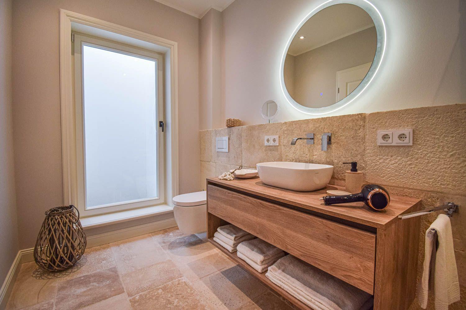 Badezimmer mit Waschtisch, Spiegel und Toilette. Fenster, Handtuchhalter und Bodenfliesen.