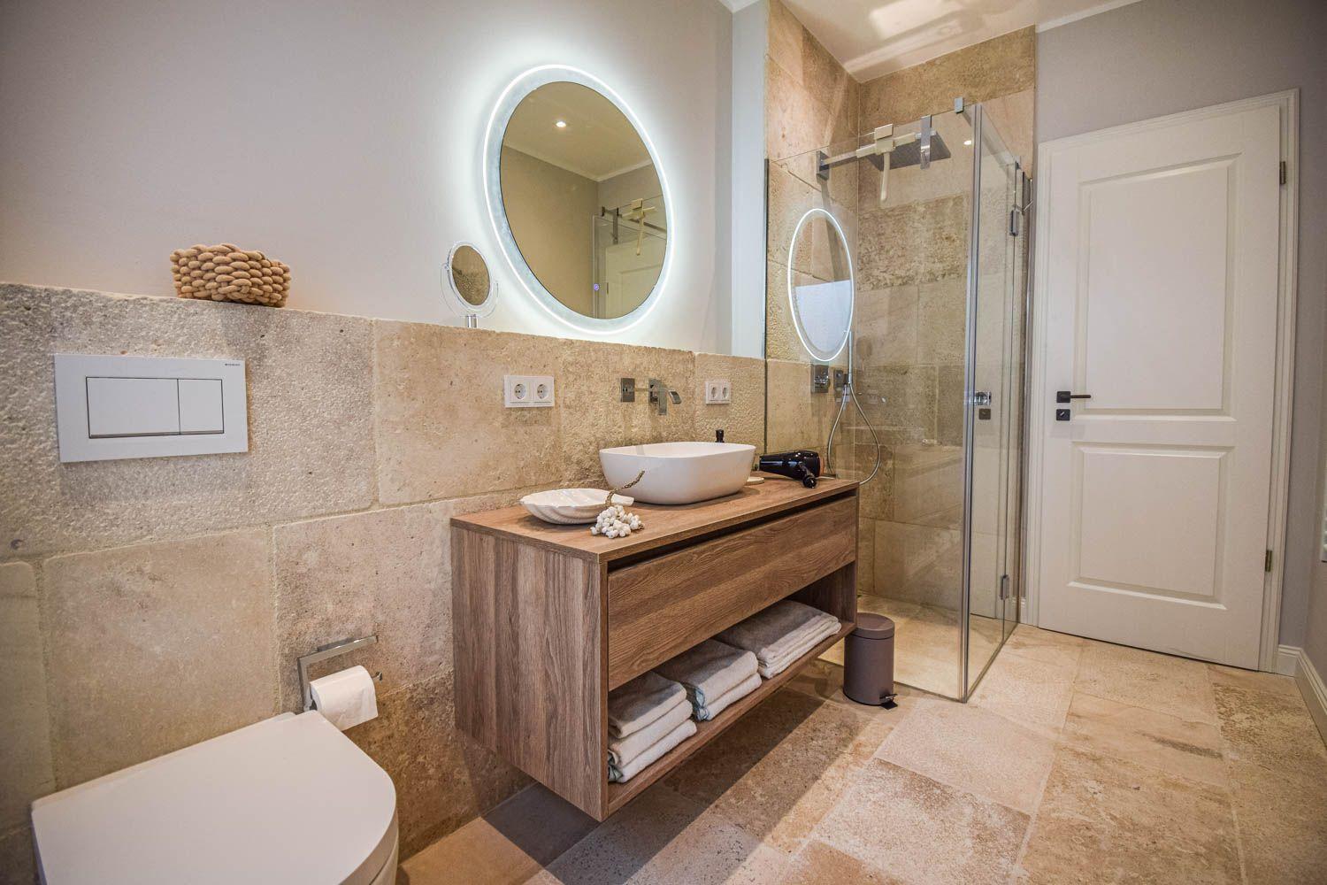 Badezimmer mit Waschbecken, Dusche und Toilette. Holzunterschrank und runder Spiegel.