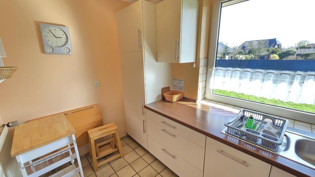 Küche mit Arbeitsplatte, Spüle und Fensterblick. Holztisch und Stuhl. Wanduhr.
